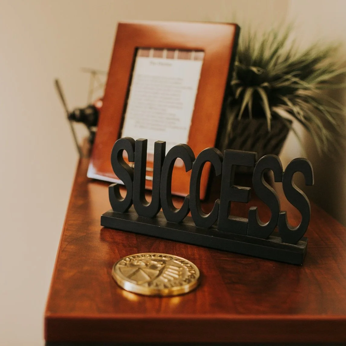 Decorative black word 'SUCCESS' on a wooden table with a gold badge and a framed document and a potted plant in the background.