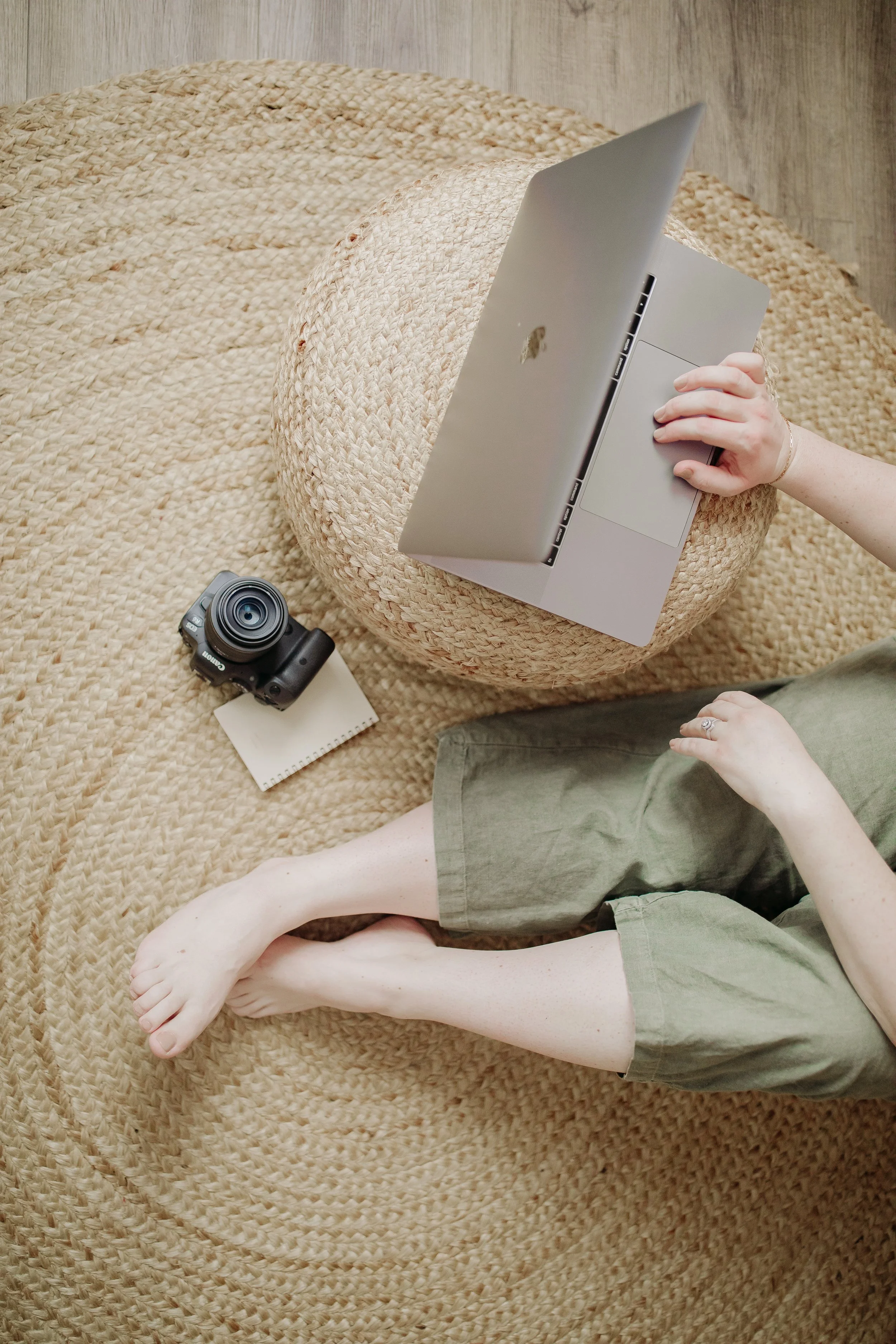 A person sitting on a woven rug with crossed legs, using a laptop, with a camera, notepad, and a large straw hat nearby.