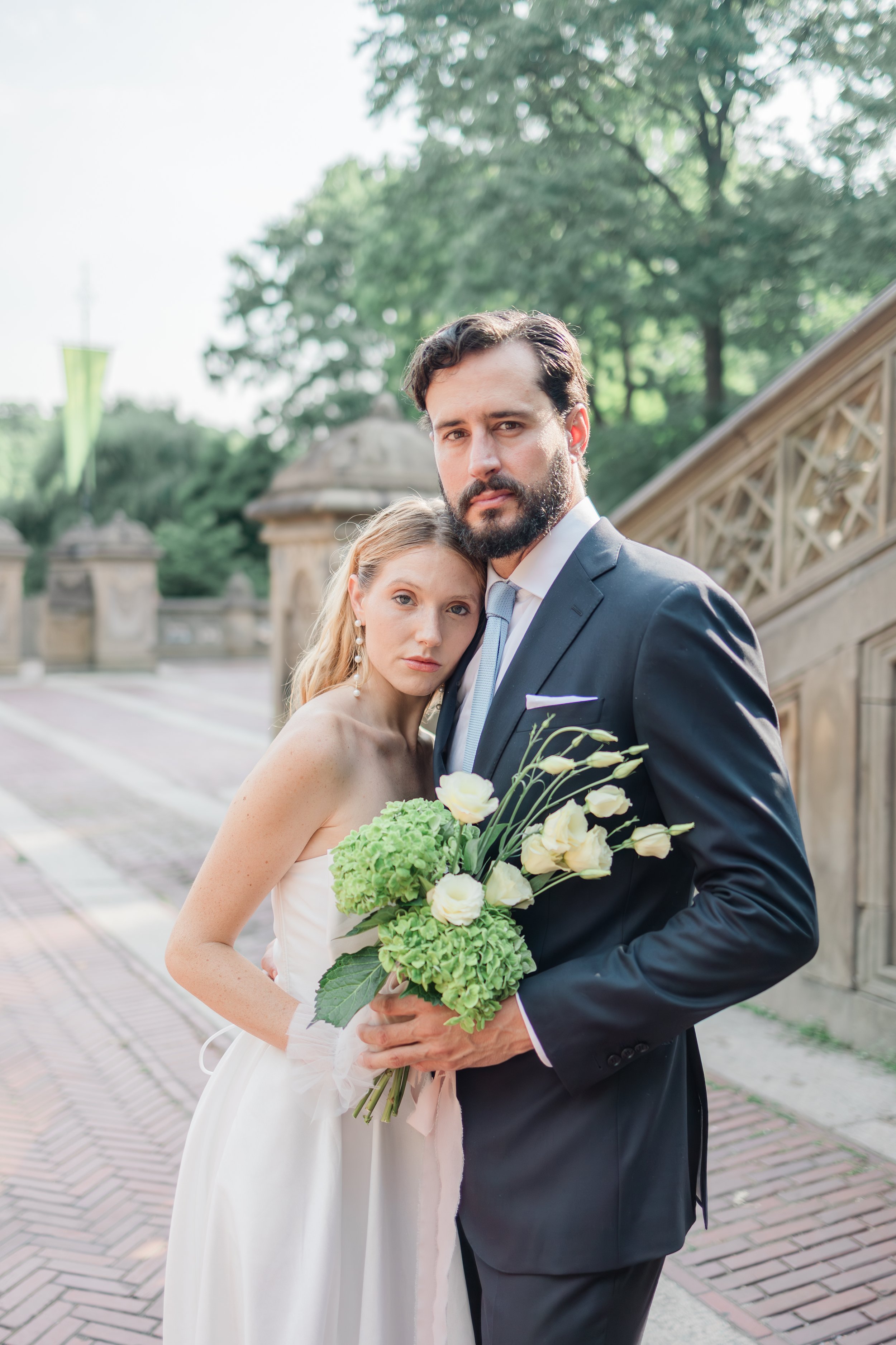 A bride and groom posing outdoors on a brick path, with green trees and a stone railing in the background. The bride holds a bouquet of green and white flowers, and they look at the camera with serious expressions.