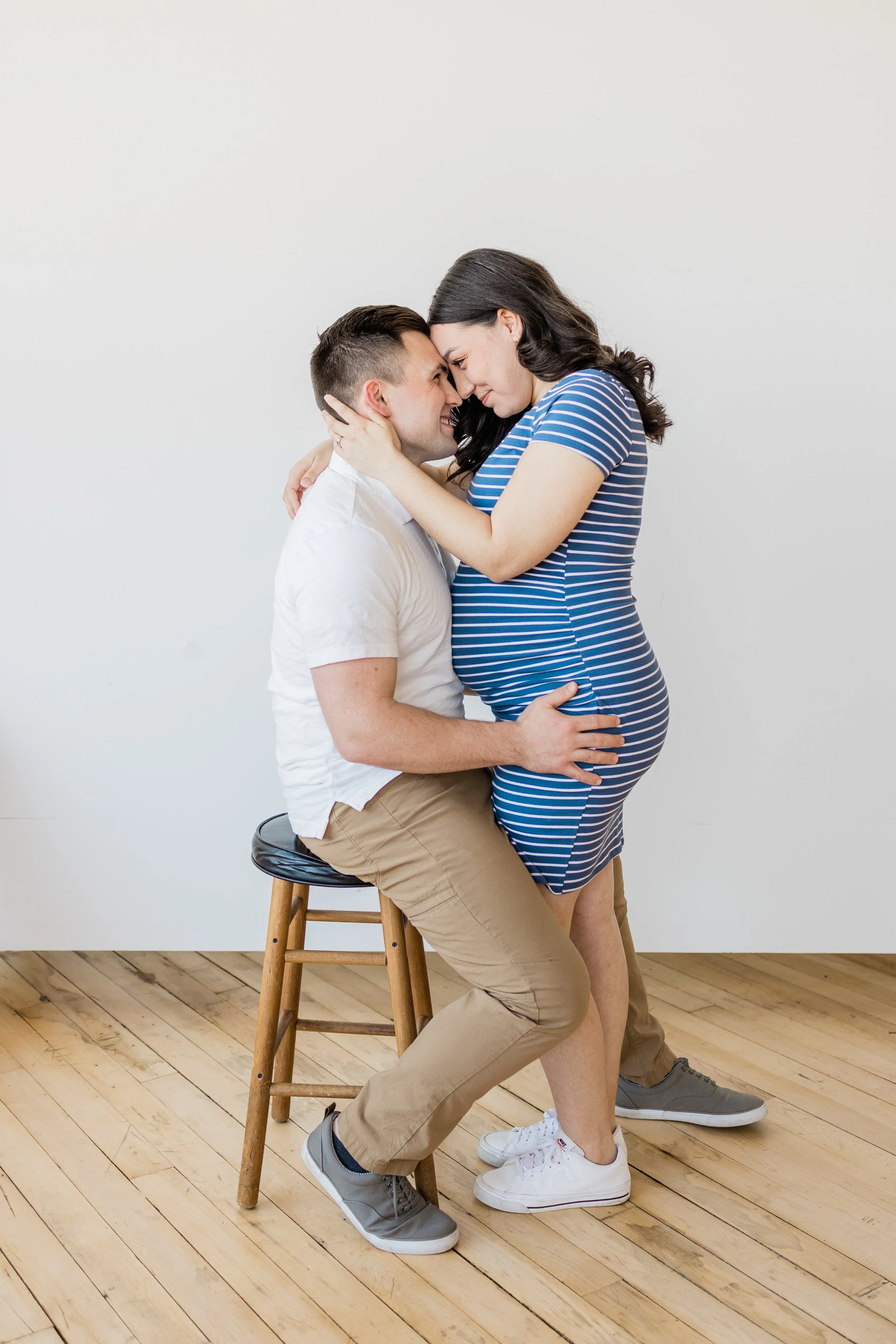 A couple sitting on a stool, with the woman visibly pregnant, touching foreheads and smiling.