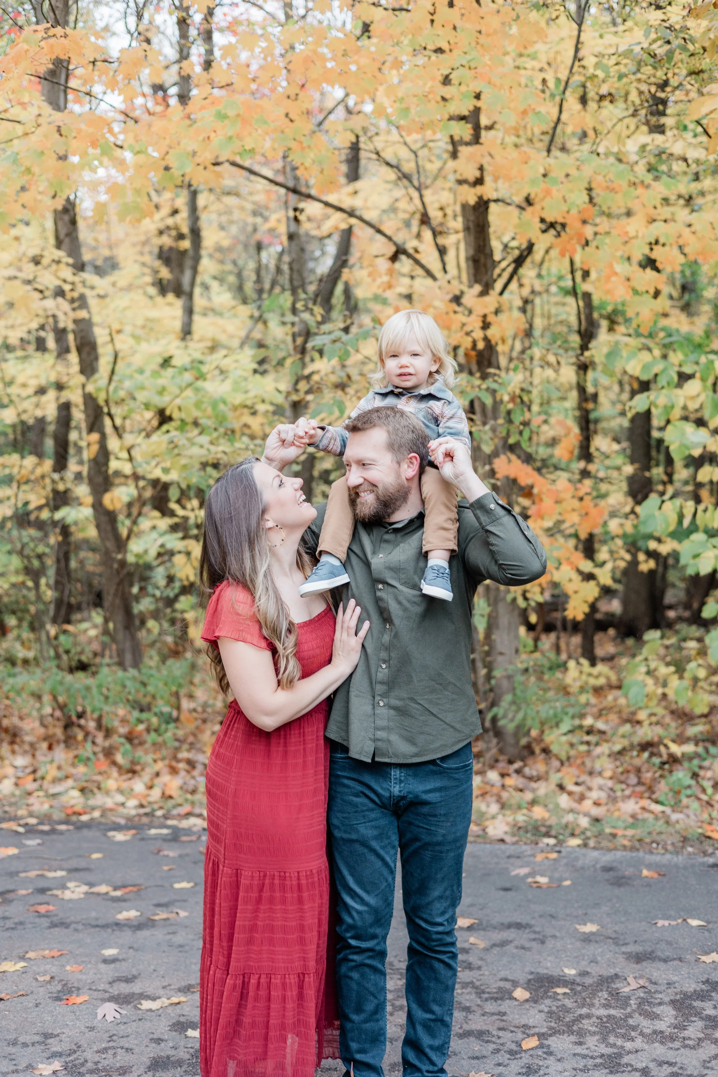 A family of three enjoying fall outdoors. The woman in a red dress and the man in a dark shirt are smiling at each other, with the woman touching the man's chest. The child, sitting on the man's shoulders, is wearing a jacket. They are surrounded by colorful autumn leaves.