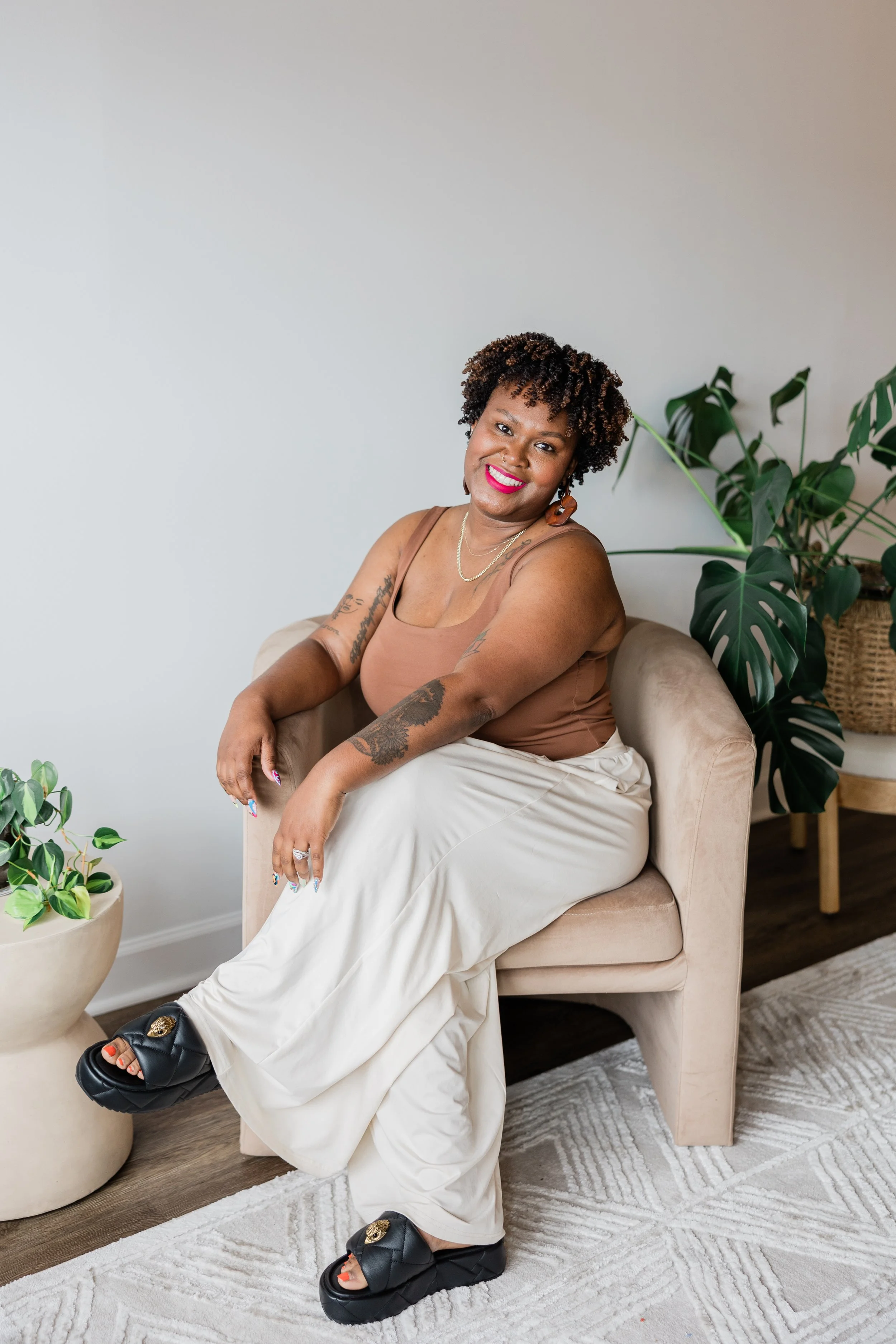 Smiling woman with short curly hair sitting on a beige armchair in a room with white walls and wooden floor, surrounded by green plants.