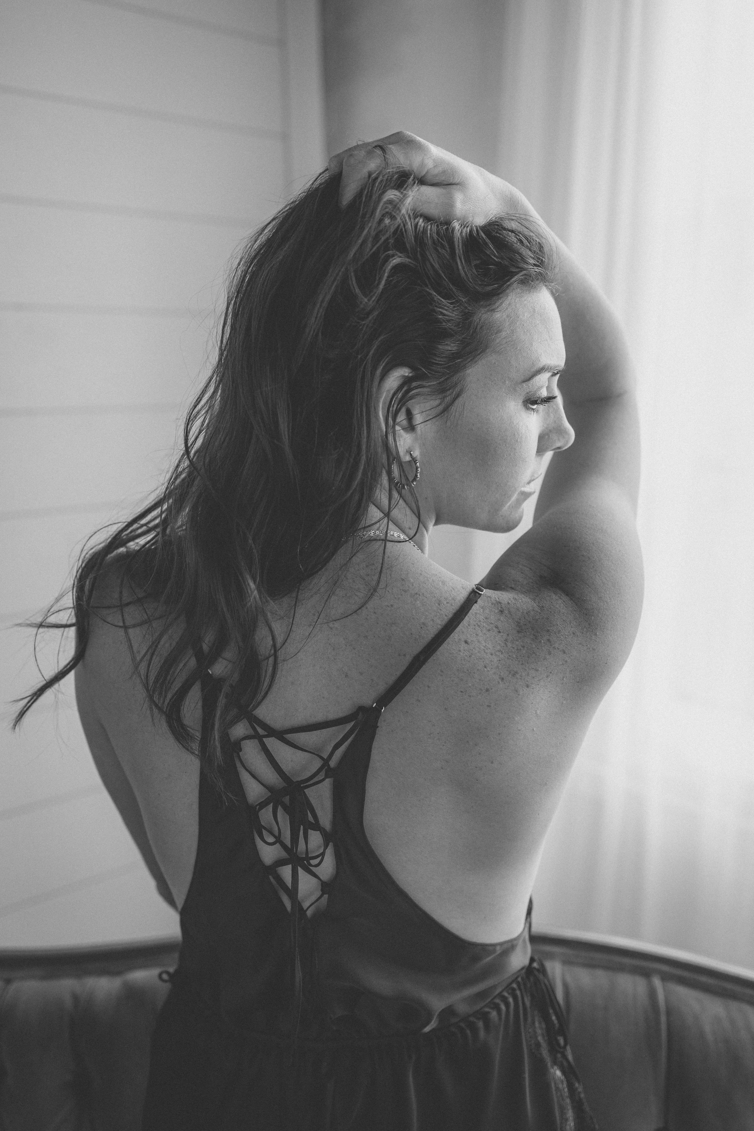 Black and white photo of a woman with long wavy hair, wearing a spaghetti strap top with laces on the back, standing near a window with her eyes closed, one hand on her head.