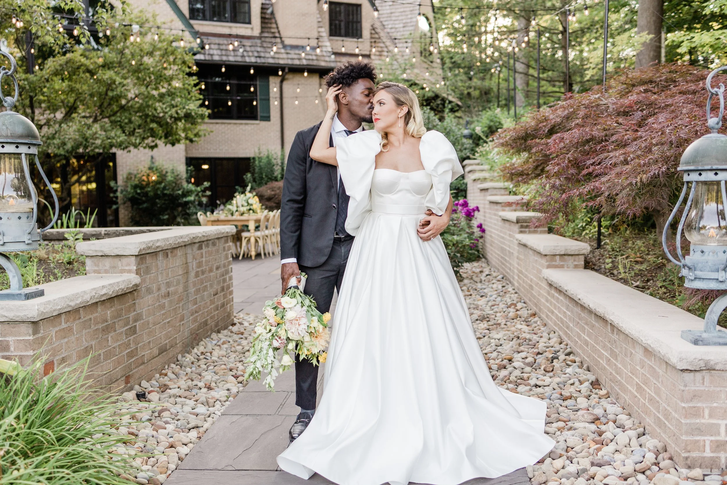 A bride and groom embrace outdoors, the bride in a white wedding gown with puffed sleeves and the groom in a black suit, holding a bouquet of flowers. The setting includes brick planters, a stone pathway, and string lights.