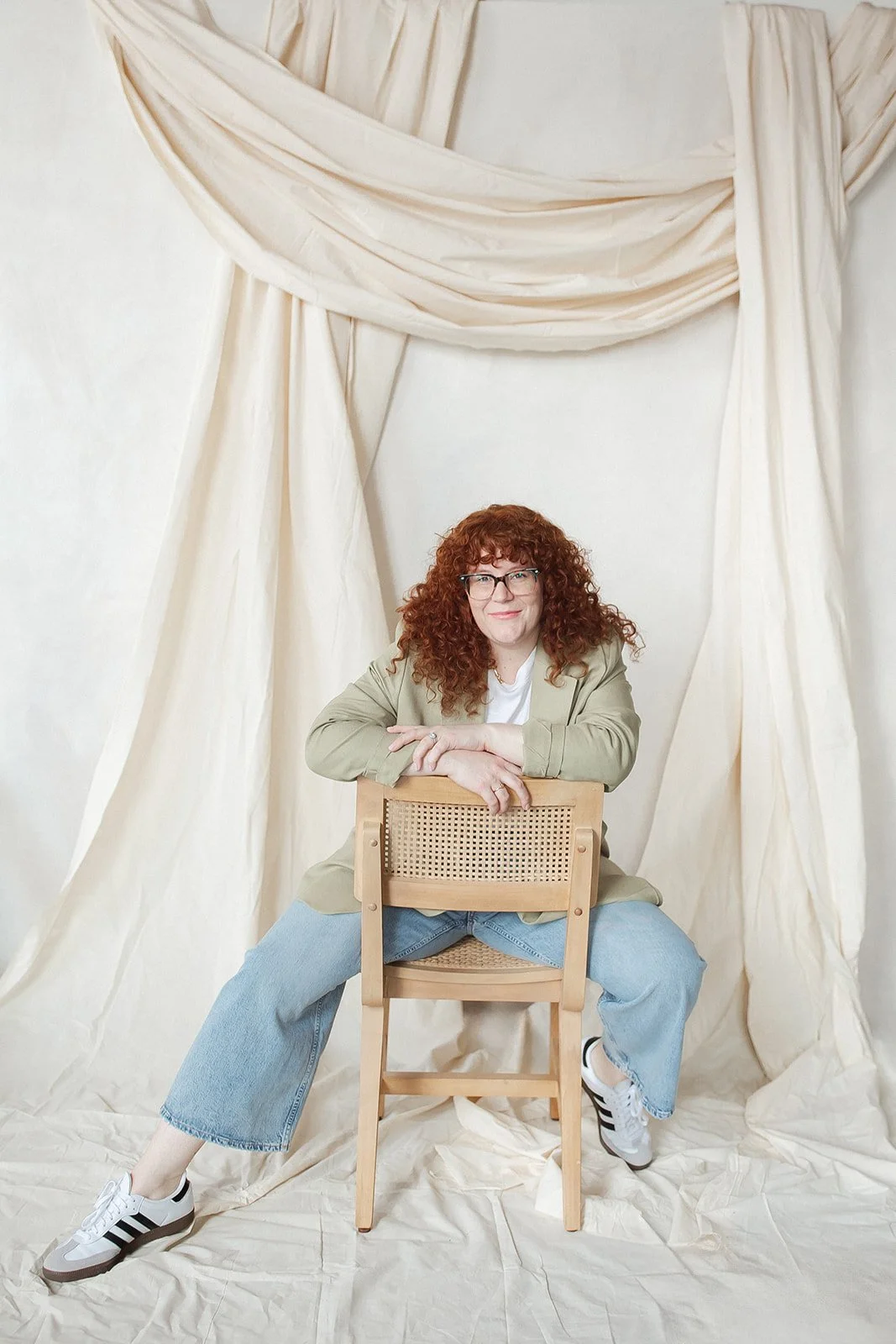 A woman with curly red hair and glasses, wearing a beige blazer, white shirt, and blue jeans, sitting on a chair with her arms resting on the backrest, against a cream-colored backdrop with draped fabric.