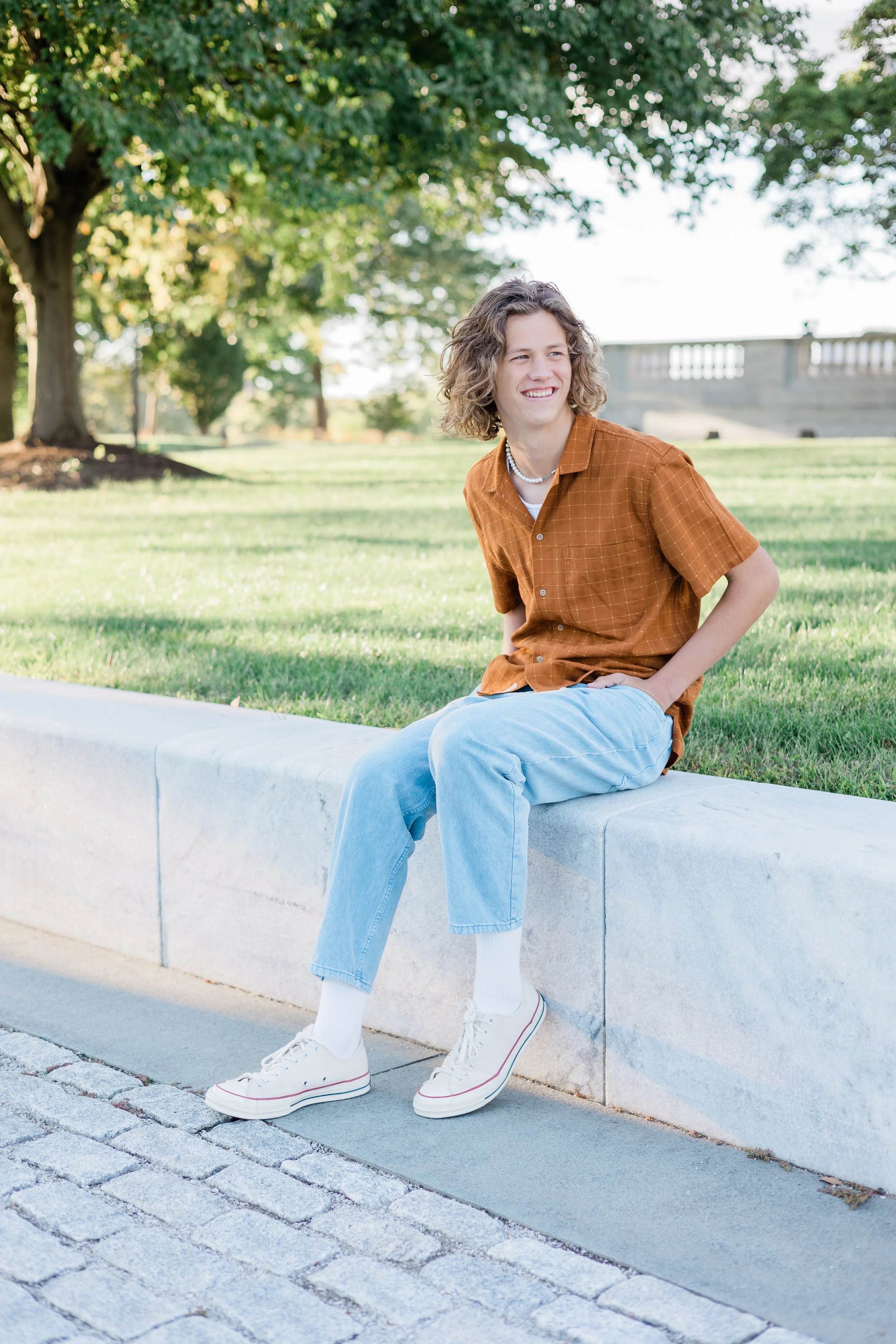 A young woman with curly hair sitting on a concrete bench outdoors, smiling, wearing an orange checkered shirt, light blue jeans, white sneakers, and a pearl necklace, with trees and grassy area in the background.