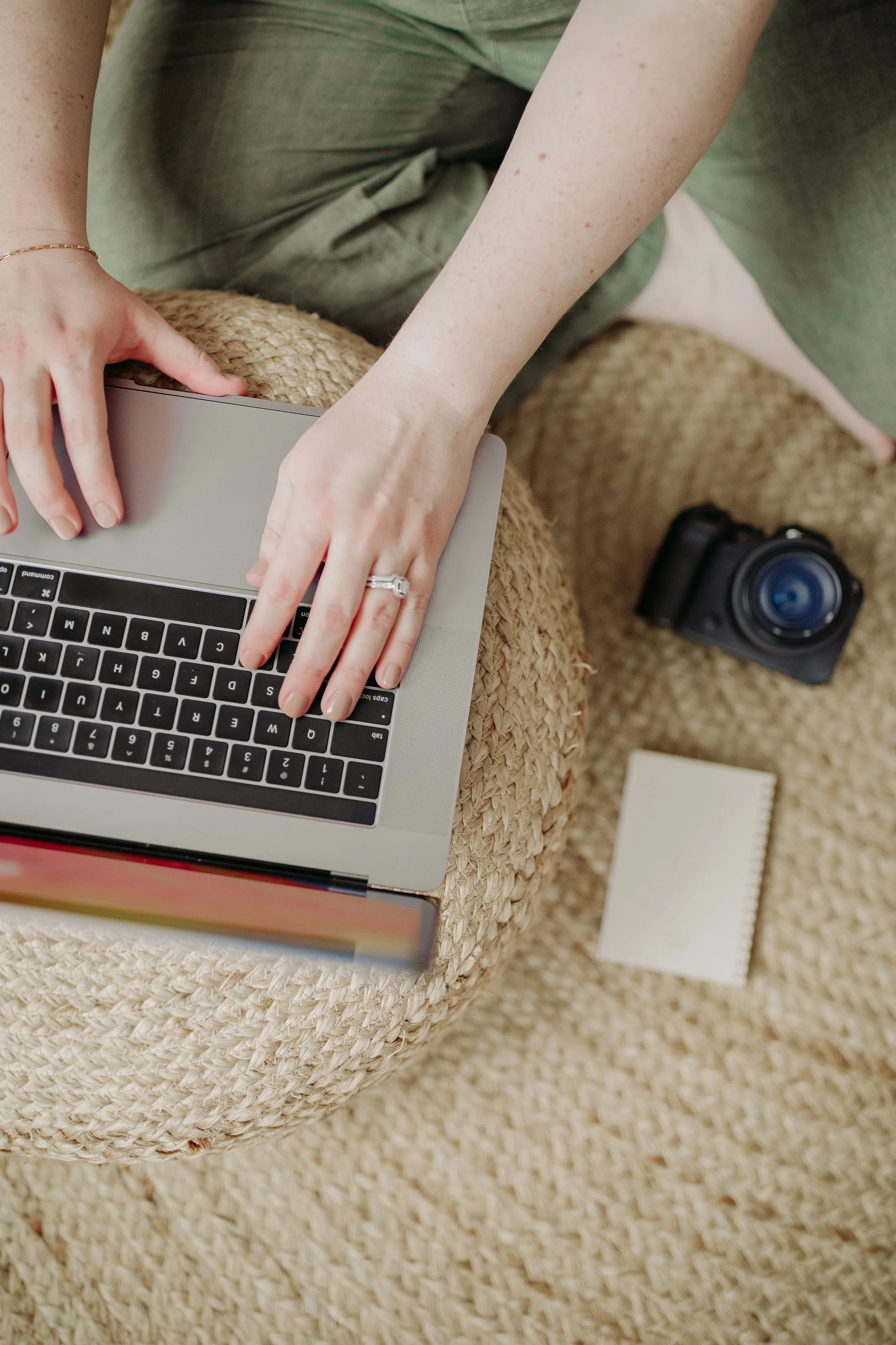 Top-down view of a person working on a laptop with a book, a camera, and a notepad on a woven stool and carpet.