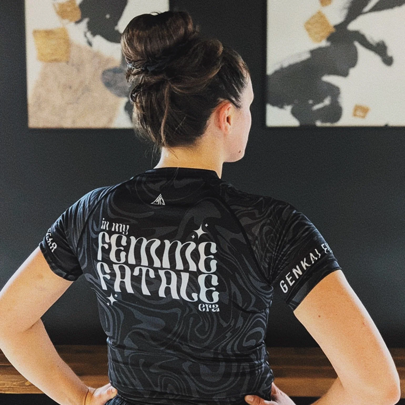 Back of woman in black athletic shirt with white text, styled hair, standing in front of abstract paintings on black wall.