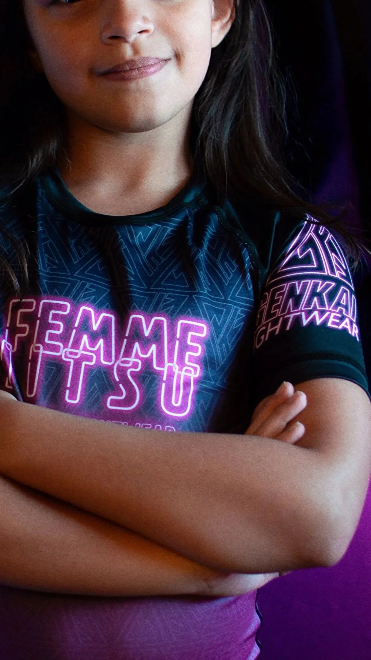Close-up of a girl with crossed arms wearing a dark sports jersey with neon pink and purple letters and geometric designs, smiling slightly at the camera.