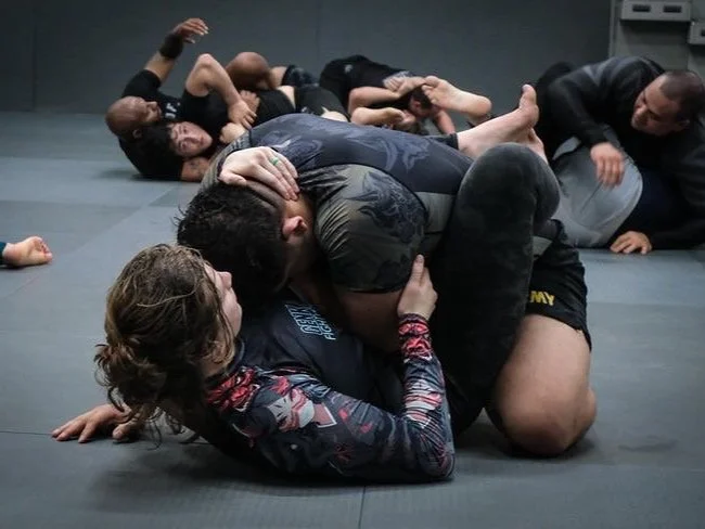 Two women practicing Brazilian jiu-jitsu on a padded mat, one woman in a black rash guard on top in a guard position, and the other in a camouflage rash guard on the bottom. Several other practitioners in black gis are training in the background.
