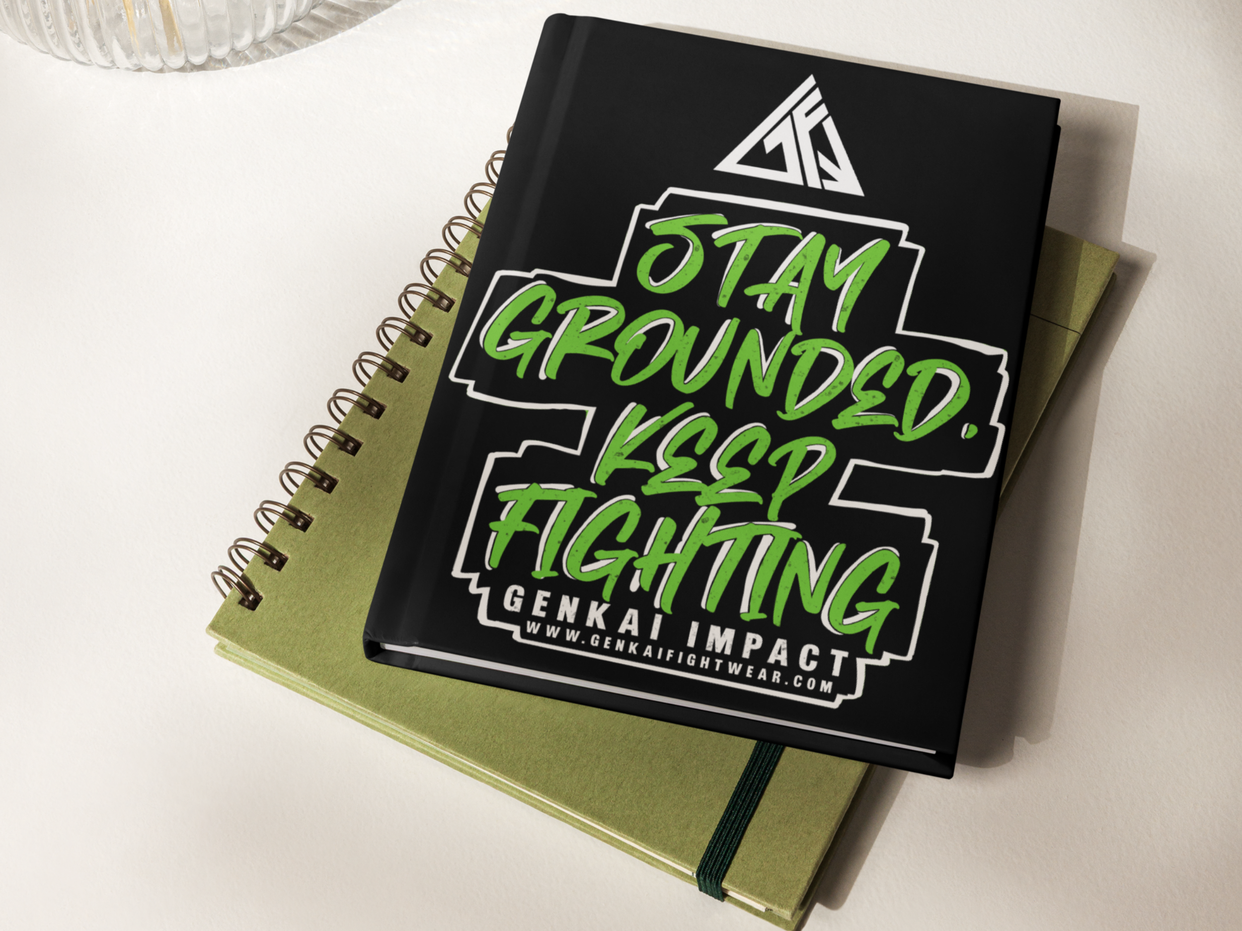 A black notebook with green and white text that says 'Stay Grounded. Keep Fighting' and a website address, placed on top of a green notebook with a black elastic band.