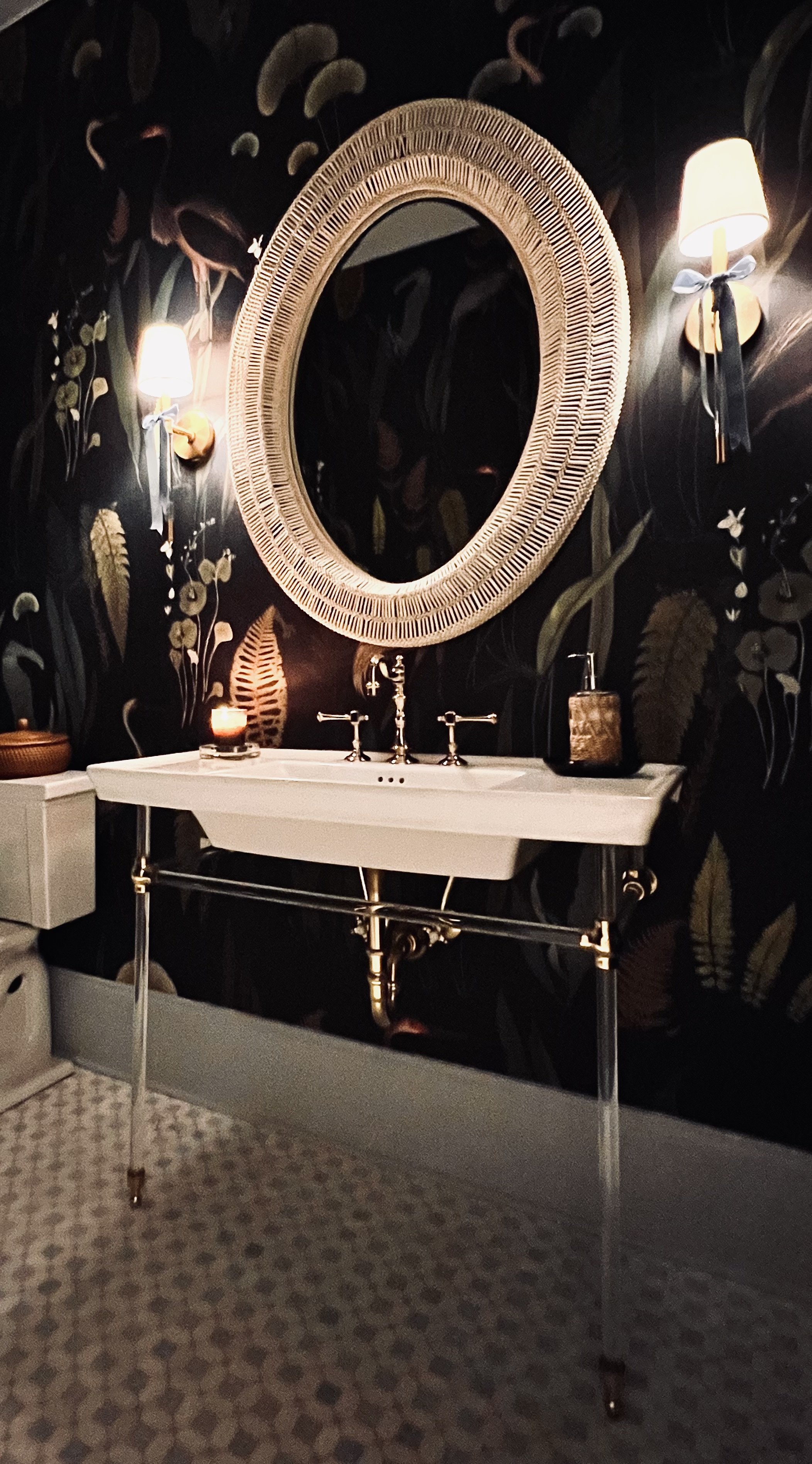 Bathroom with ornate mirror, dual wall sconces, floral wallpaper, white sink and countertop, and decorative candle.