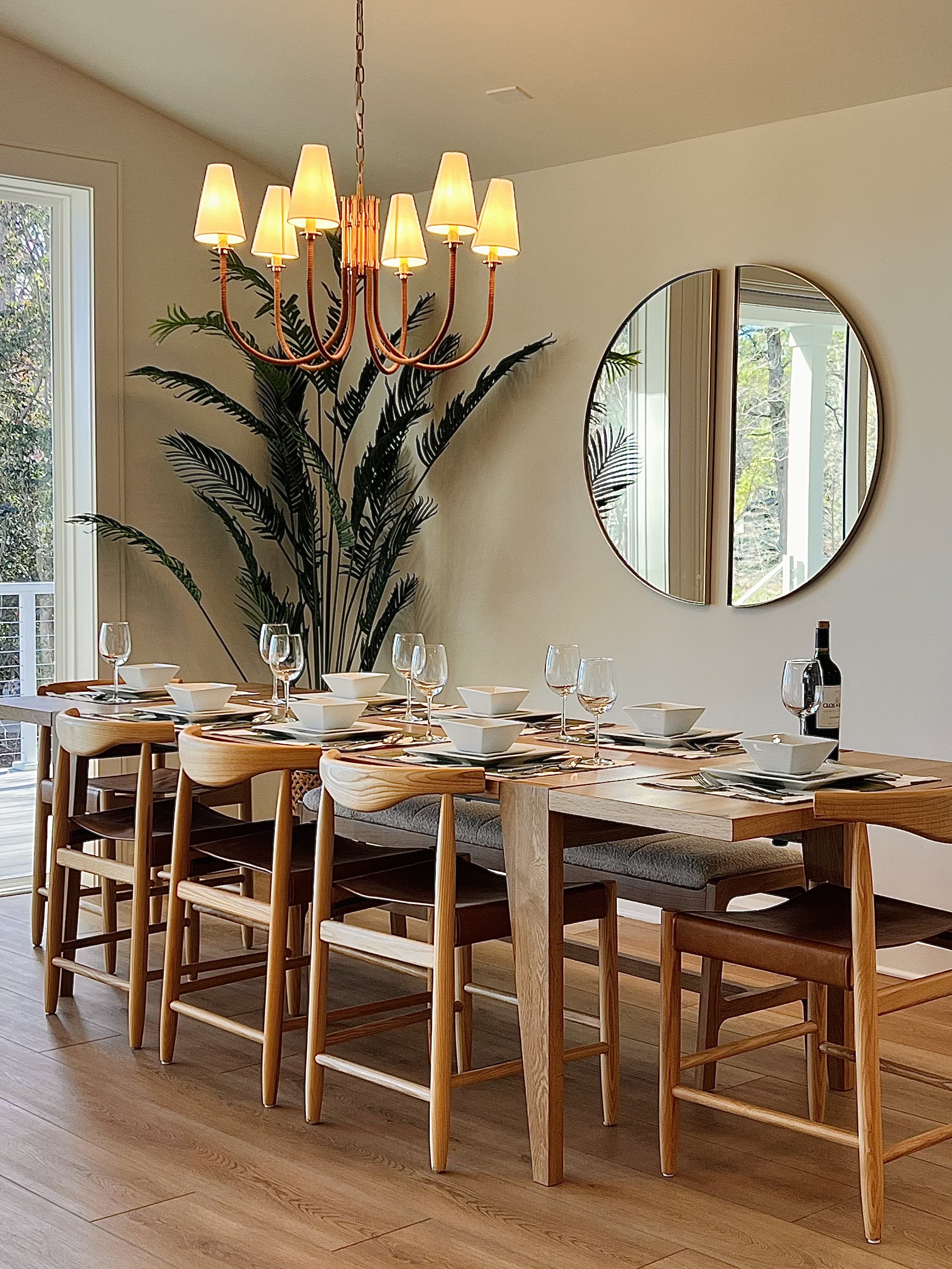 Modern dining room setup with wooden table, chairs, and a chandelier. The table is set with plates, glasses, and a bottle of wine. A large round mirror and a potted plant are in the background.