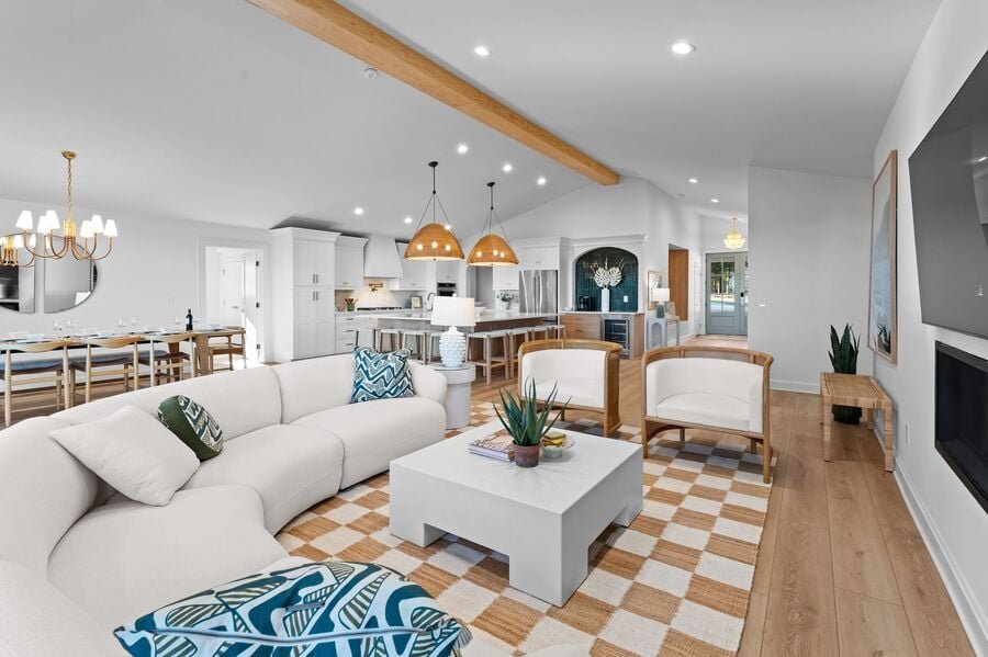 Modern living room with white furniture, wooden accents, and open layout. Features a white sectional sofa, patterned cushions, a square coffee table, and a checkered rug. The space includes a kitchen with pendant lights and a dining area with a chand