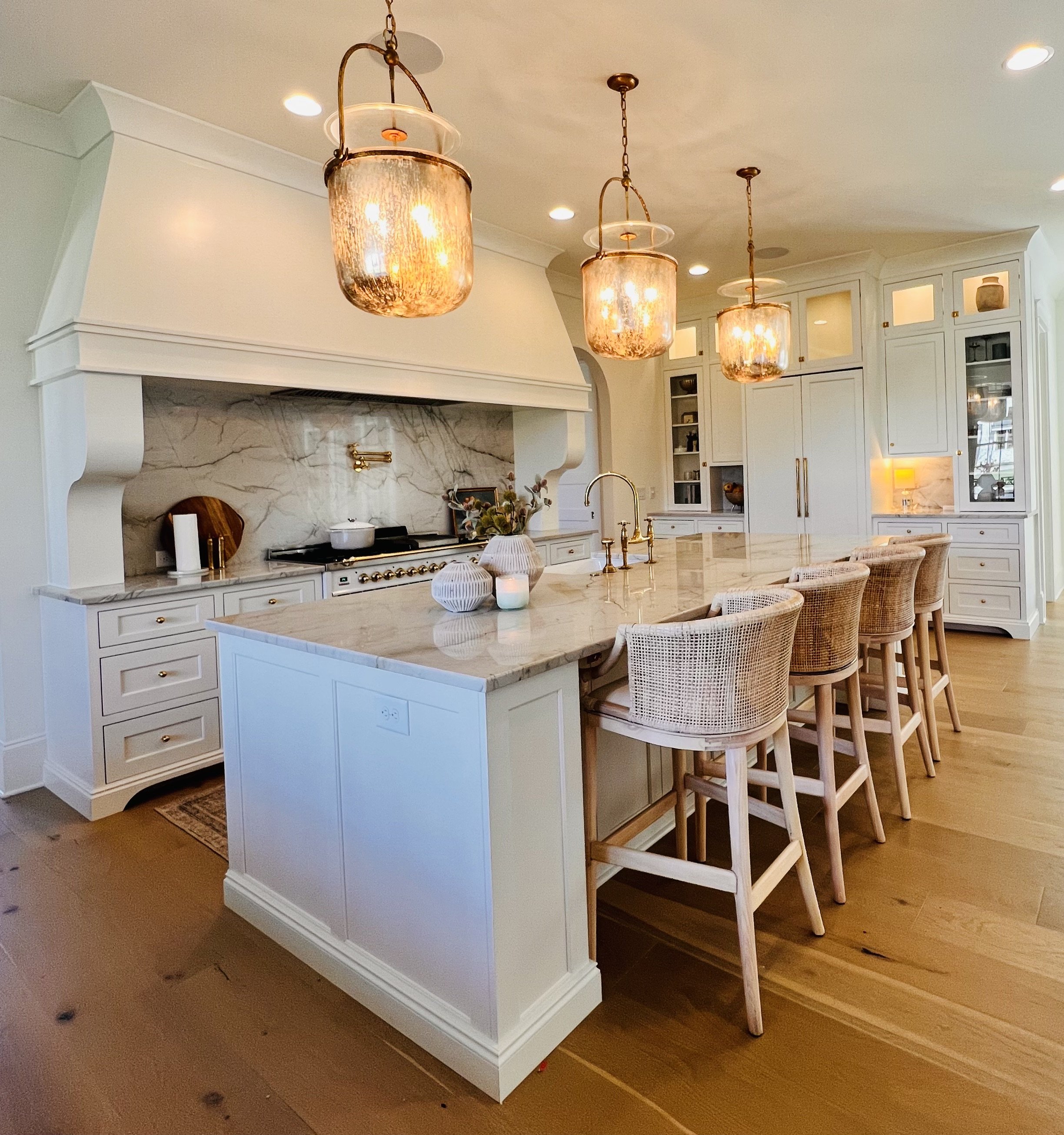Modern kitchen with marble countertops, wicker bar stools at an island, pendant lights, white cabinetry, and a large hood over the stove.