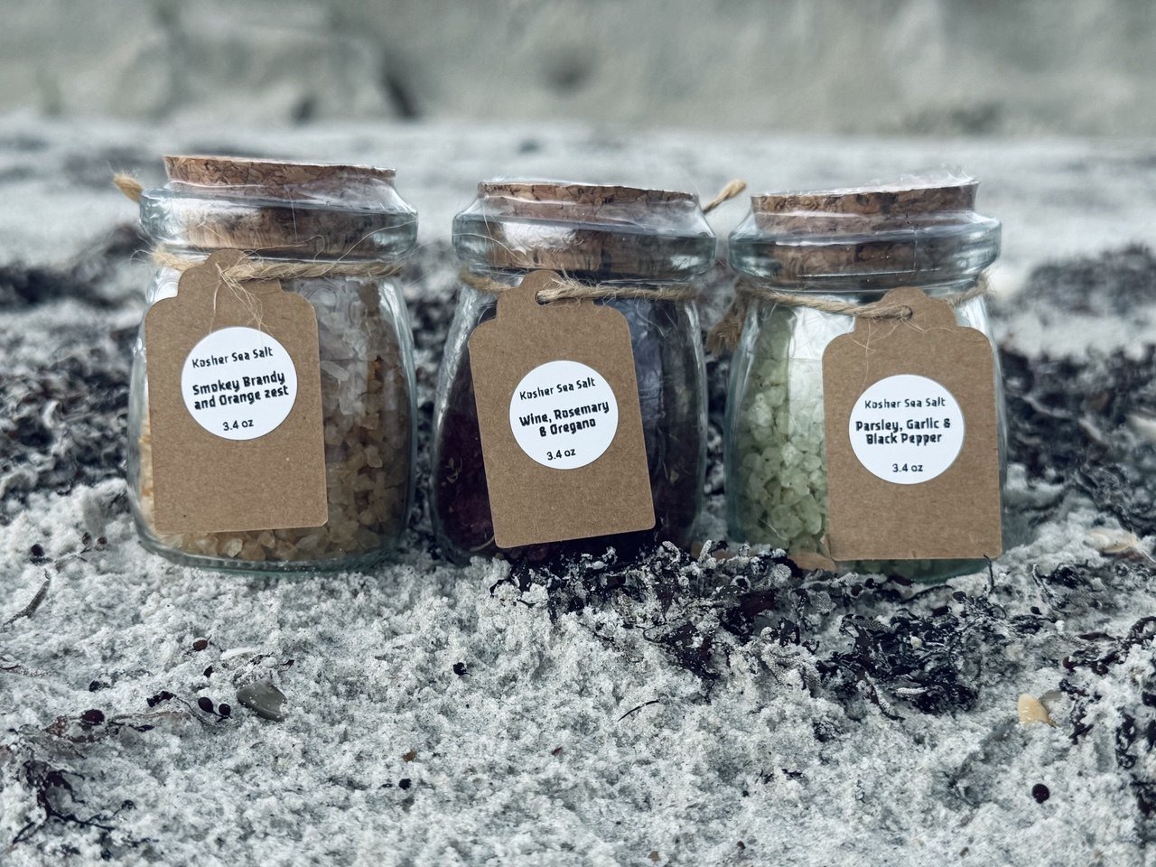 Three small glass jars of flavored sea salts with kraft paper tags, placed on sandy ground. The flavors are smoked brisket and orange zest, wine rosemary and oregano, and parsley garlic and black pepper.