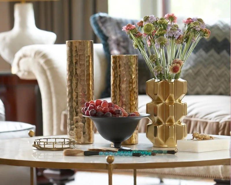 A decorative living room table arrangement with pink and purple flowers in a gold geometric vase, two hammered gold candles, a bowl of grapes, a small white tray with a gold bracelet, a makeup brush, and a turquoise beaded necklace, with a beige upholstered sofa and patterned cushions in the background.