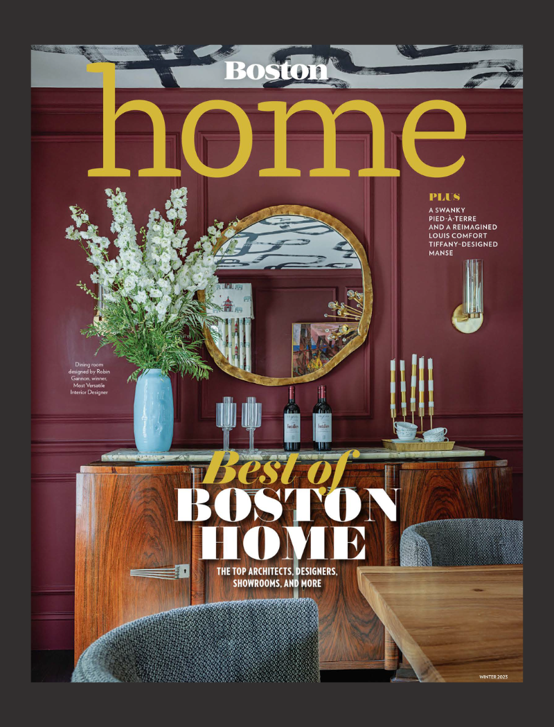 Interior of a stylish dining room with a maroon wall, a large round mirror, a wooden sideboard, a tall vase with white flowers, wine bottles, glasses, and decorative candles.