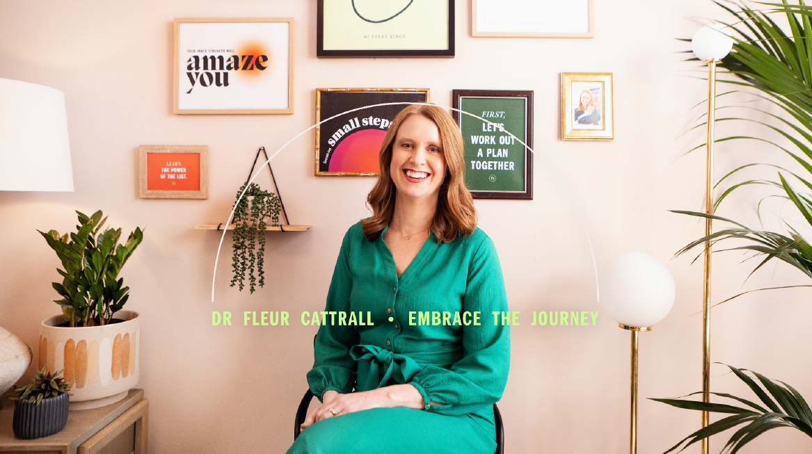 A woman with shoulder-length brown hair wearing a green blouse sitting in a room with framed artwork and plants on the wall and surrounding her.