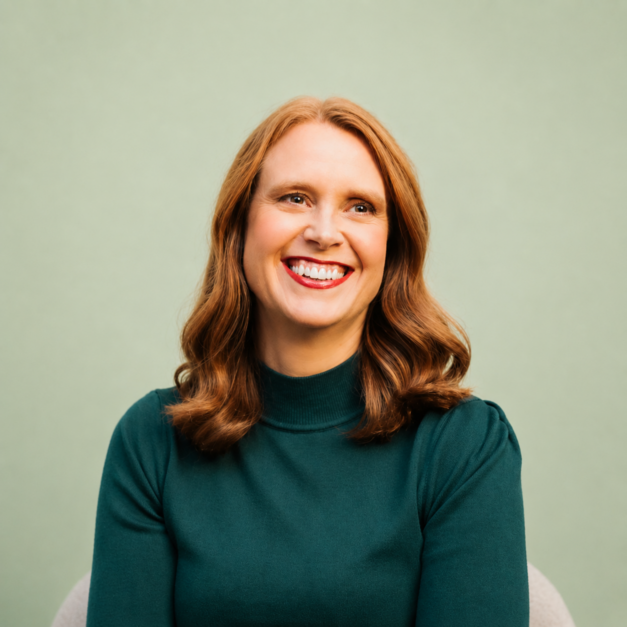 A woman with red hair, smiling, wearing a dark green turtleneck sweater, sitting against a light green background.