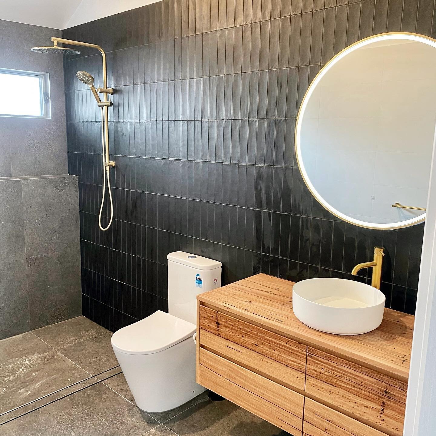 Black matte finish vertical subway feature wall, gold tapware and a touch of timber to soften this bathroom ✅
