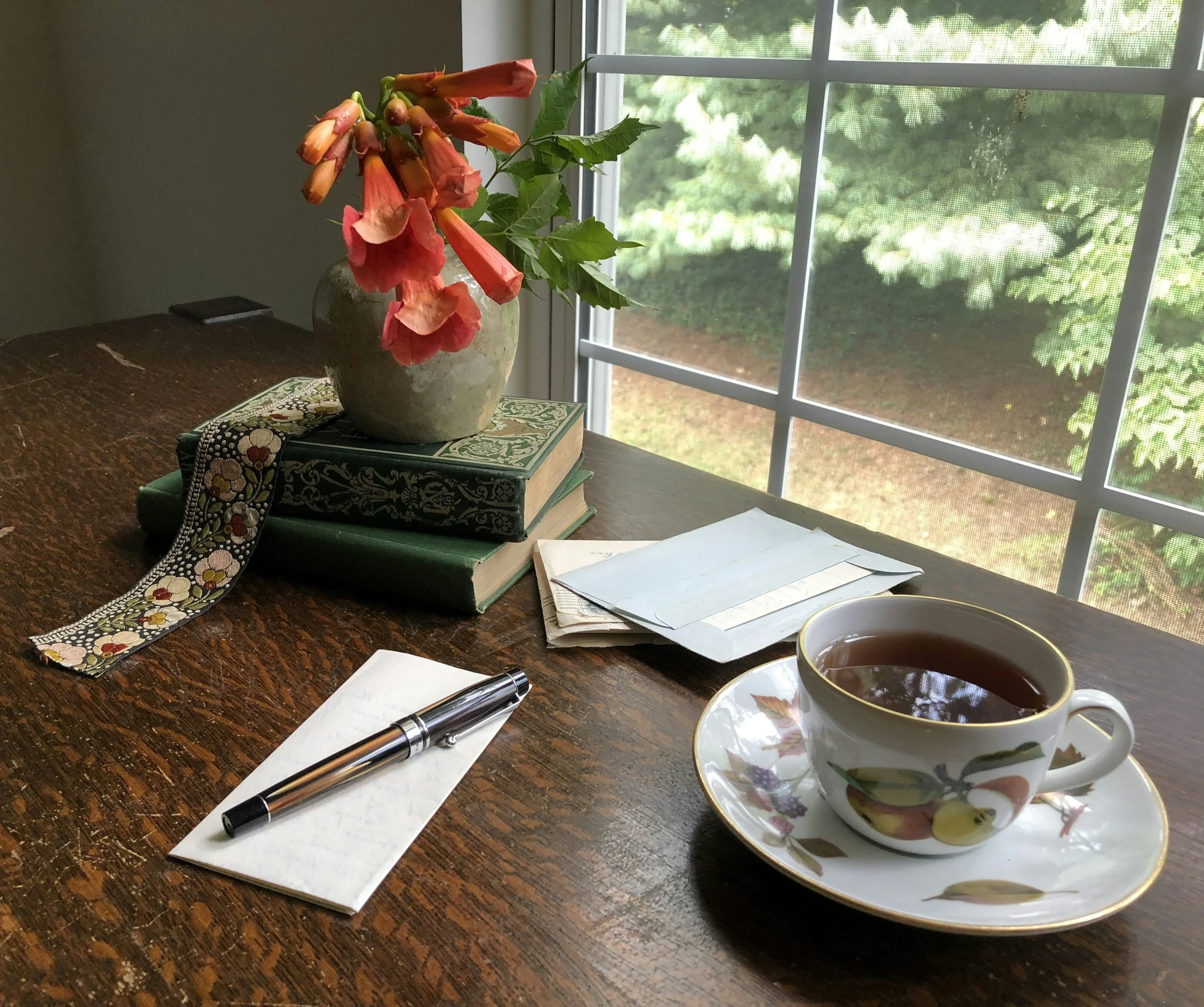 A wooden table near a window with a flower vase, books, papers, a pen, and a cup of tea.
