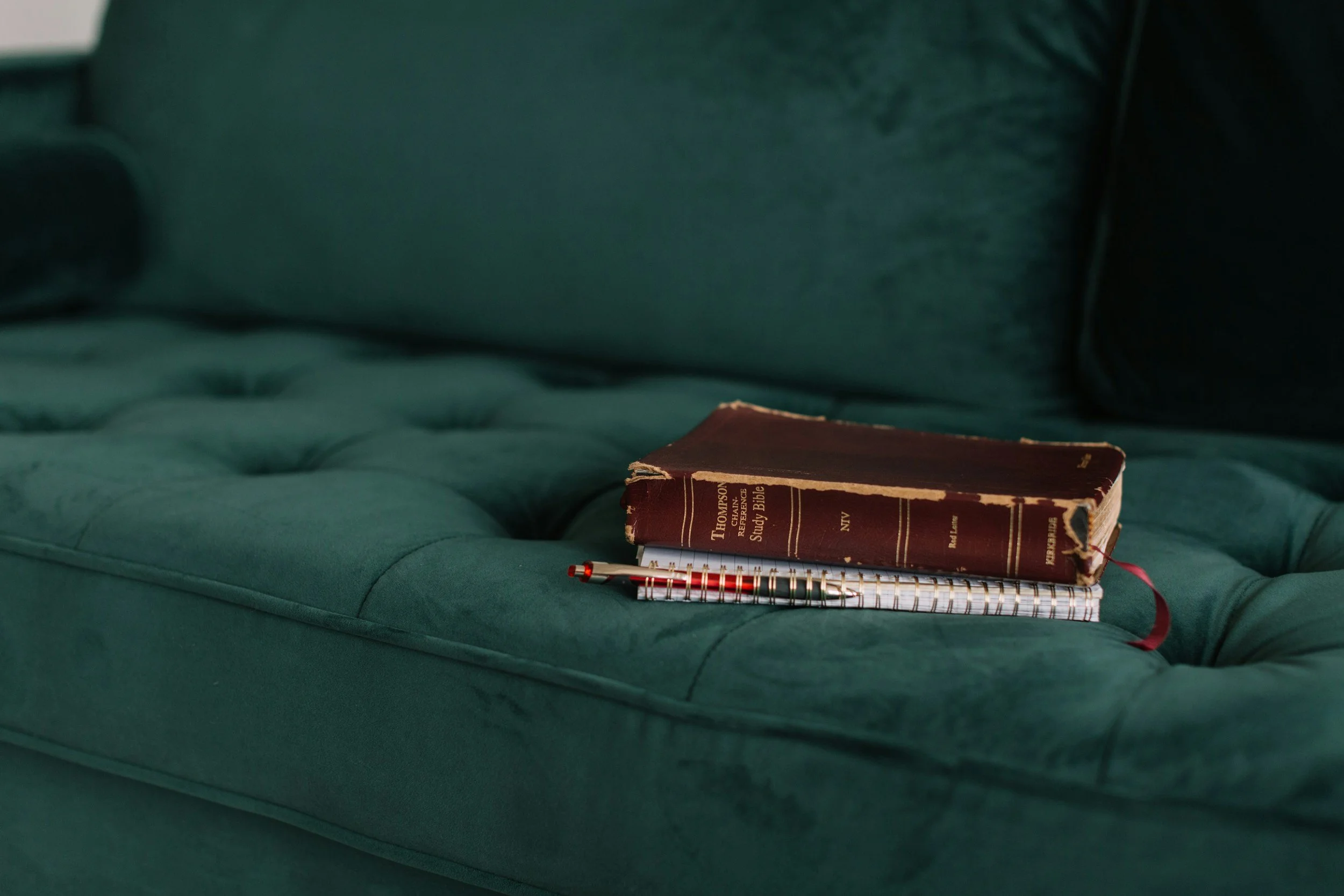 A worn Bible resting on a teal tufted sofa, with a spiral notebook and pen placed on top of it.