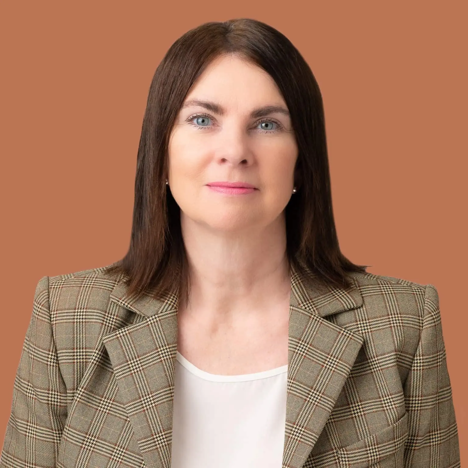 A woman with straight, shoulder-length brown hair and blue eyes, wearing a plaid blazer and a white blouse, standing against a solid brown background.