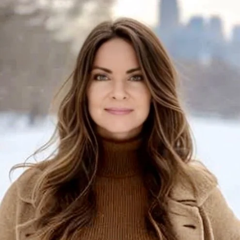 A woman with long brown hair, wearing a brown turtleneck sweater and a tan coat, standing outdoors in a snowy environment with blurred trees and buildings in the background.