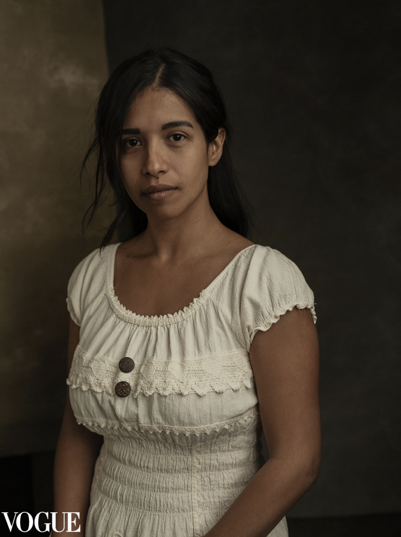 A young woman with long dark hair and medium skin tone posing indoors, wearing a cream-colored dress with decorative buttons and lace details, against a dark background.
