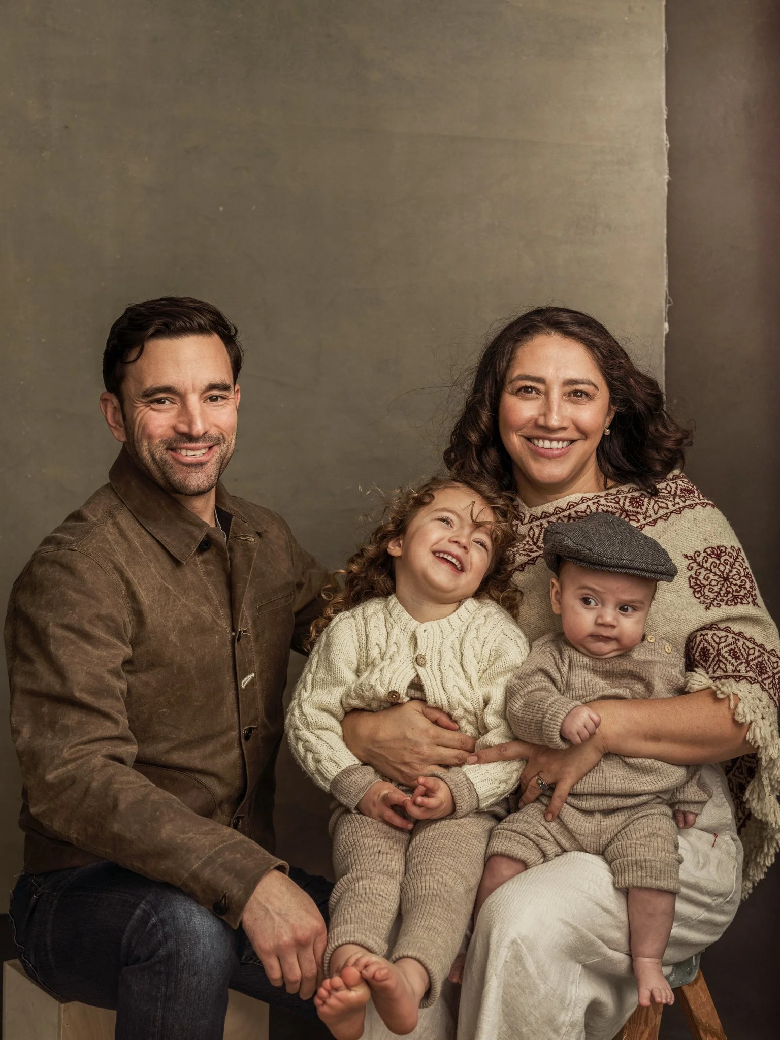 A smiling family of four sitting together in front of a simple background, with two adults and two children.