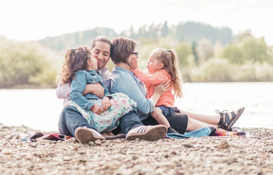A family of four sitting on a rocky beach near a body of water, engaging in a joyful moment. The father and mother sit close, with two young girls, one on each side, all smiling and interacting with each other.