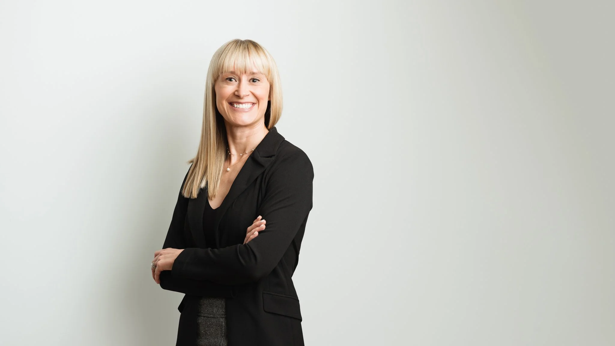 A smiling woman with long blonde hair and bangs, wearing a black blazer, standing with arms crossed against a plain light gray background.