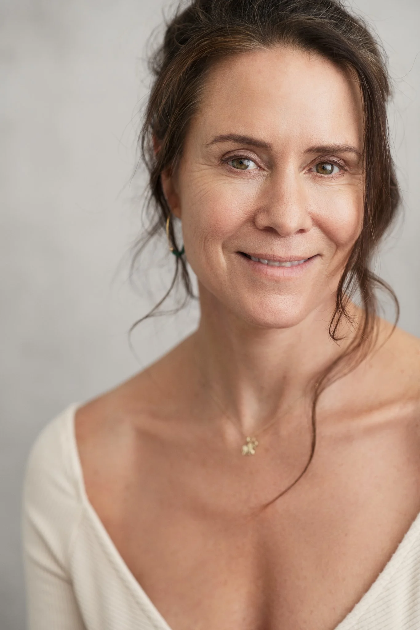 Close-up of a woman with brown hair, wearing a white top, and smiling softly. She has gold hoop earrings and a delicate necklace with a four-leaf clover pendant.
