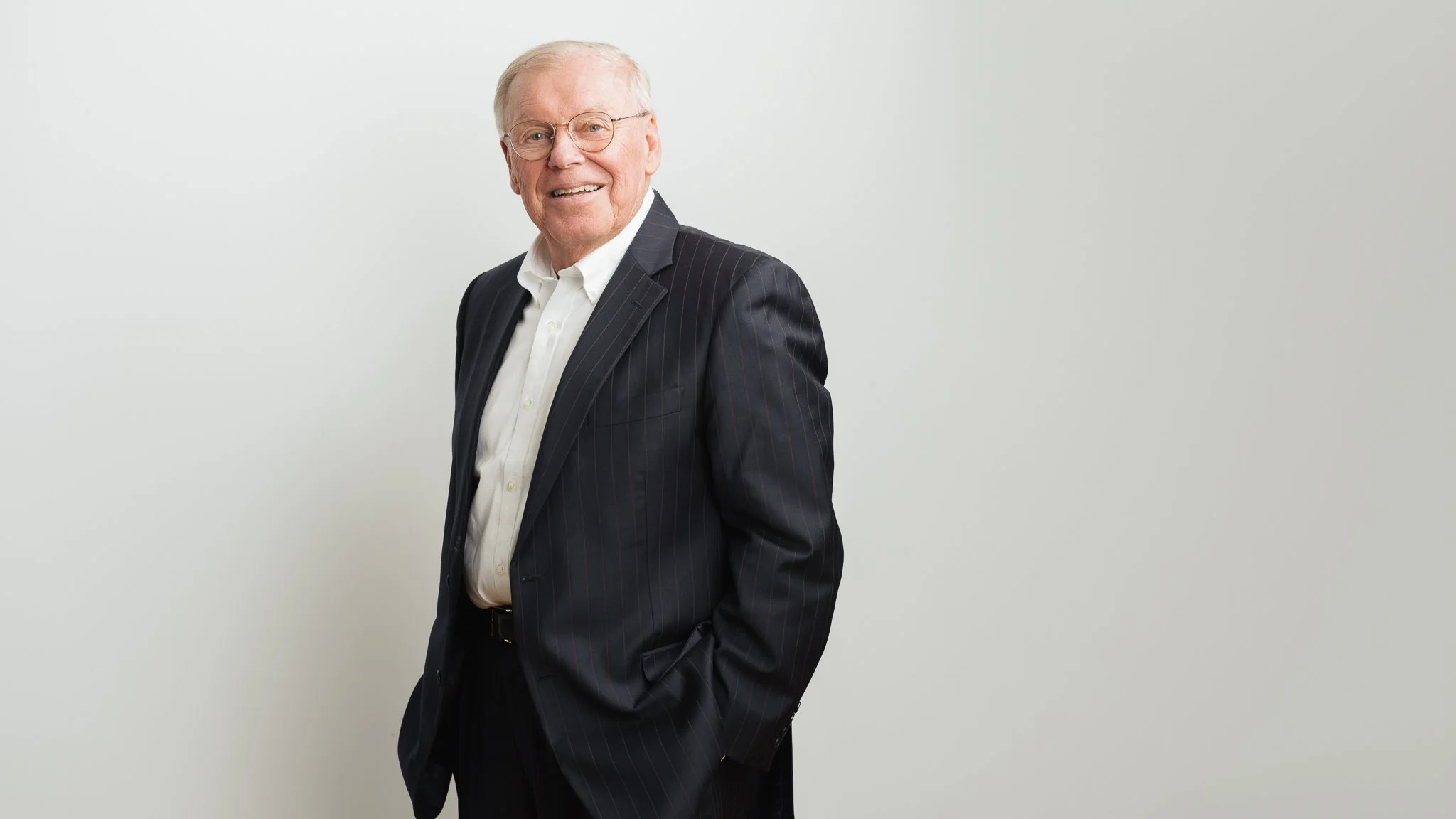 An elderly man wearing a black pinstripe suit, white dress shirt, and glasses, smiling and standing against a plain white background.