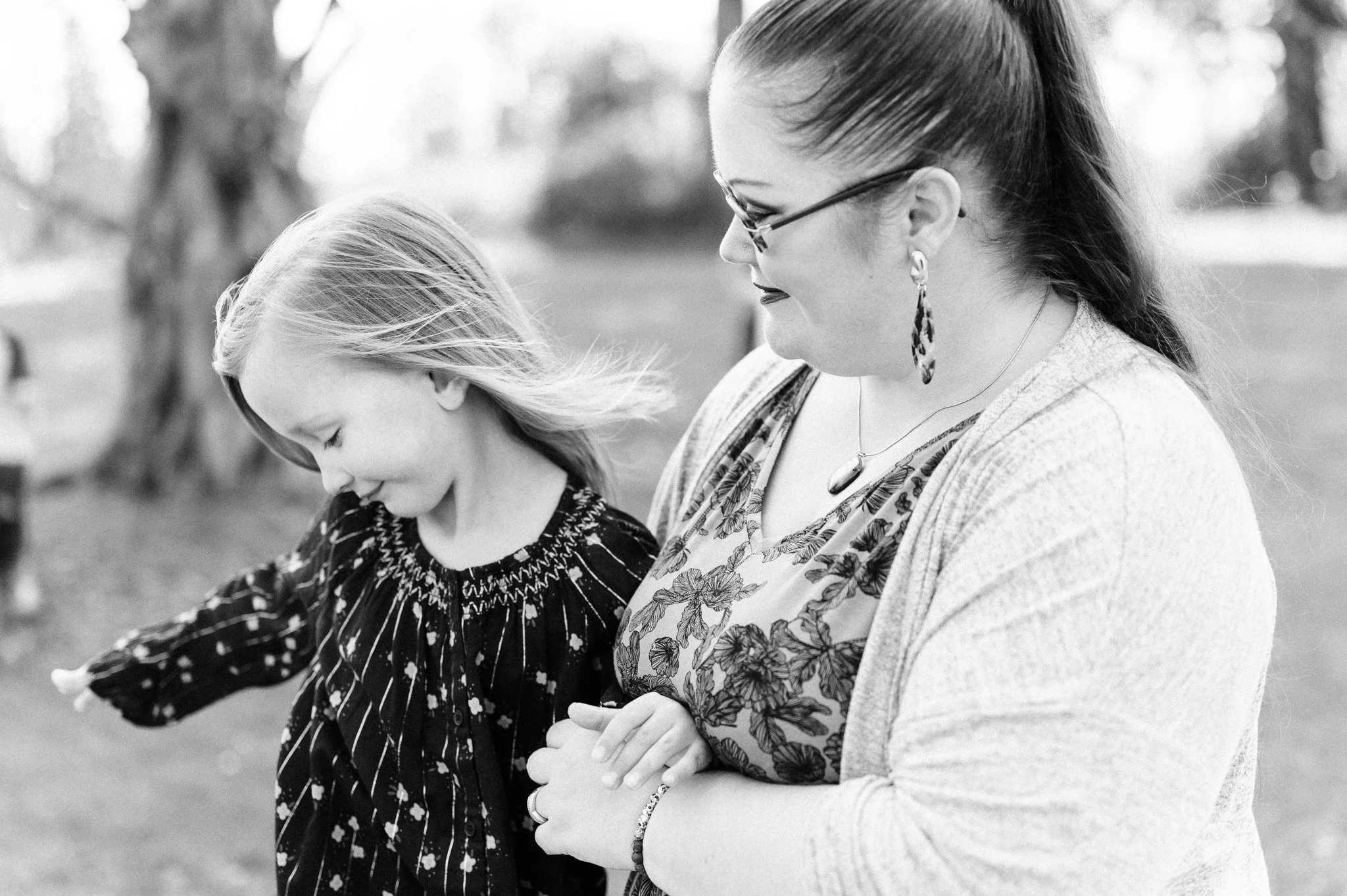 A woman and a young girl smiling outdoors in a park.
