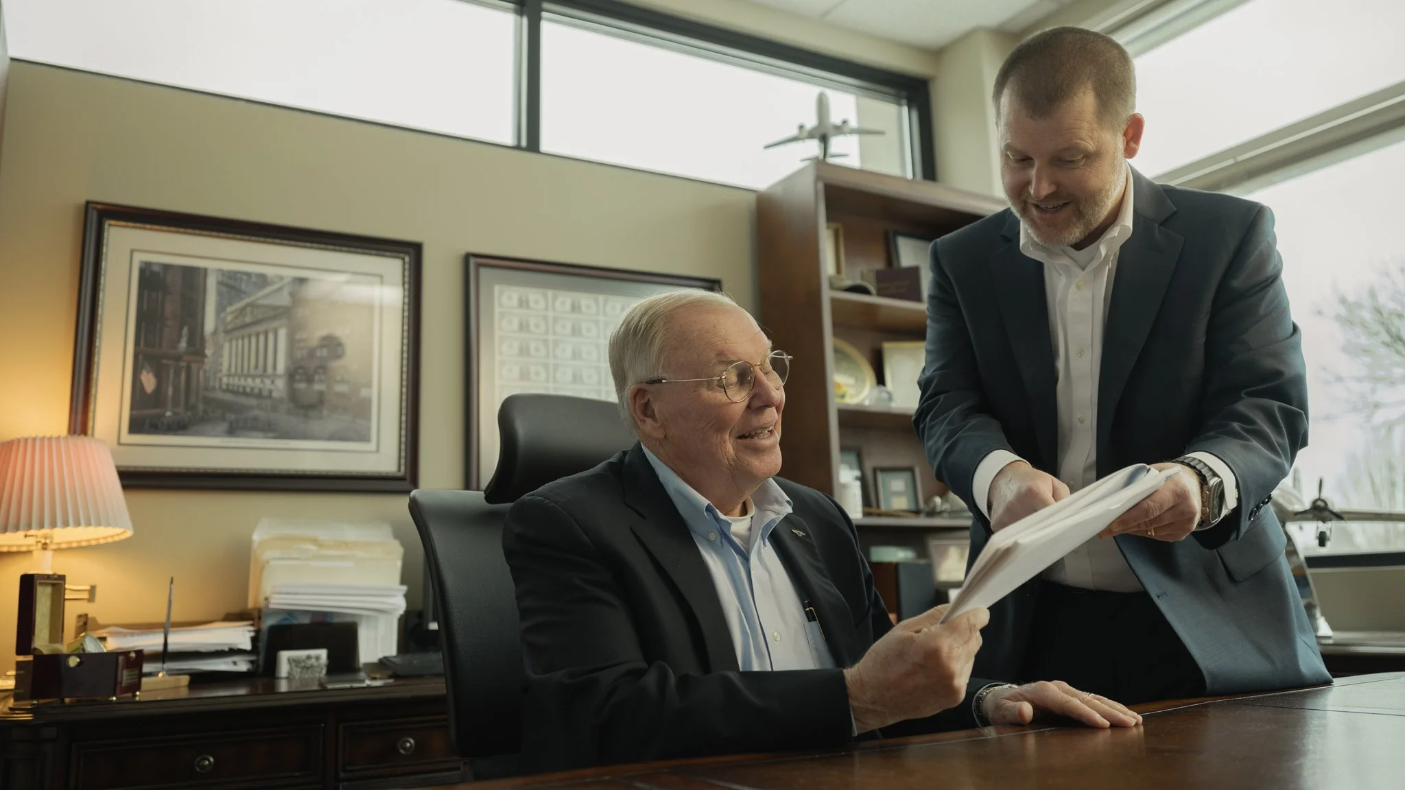 Two men in business attire in an office. One seated and the other standing, looking at documents. The office has framed pictures, a bookshelf, and a window with a model airplane on top.