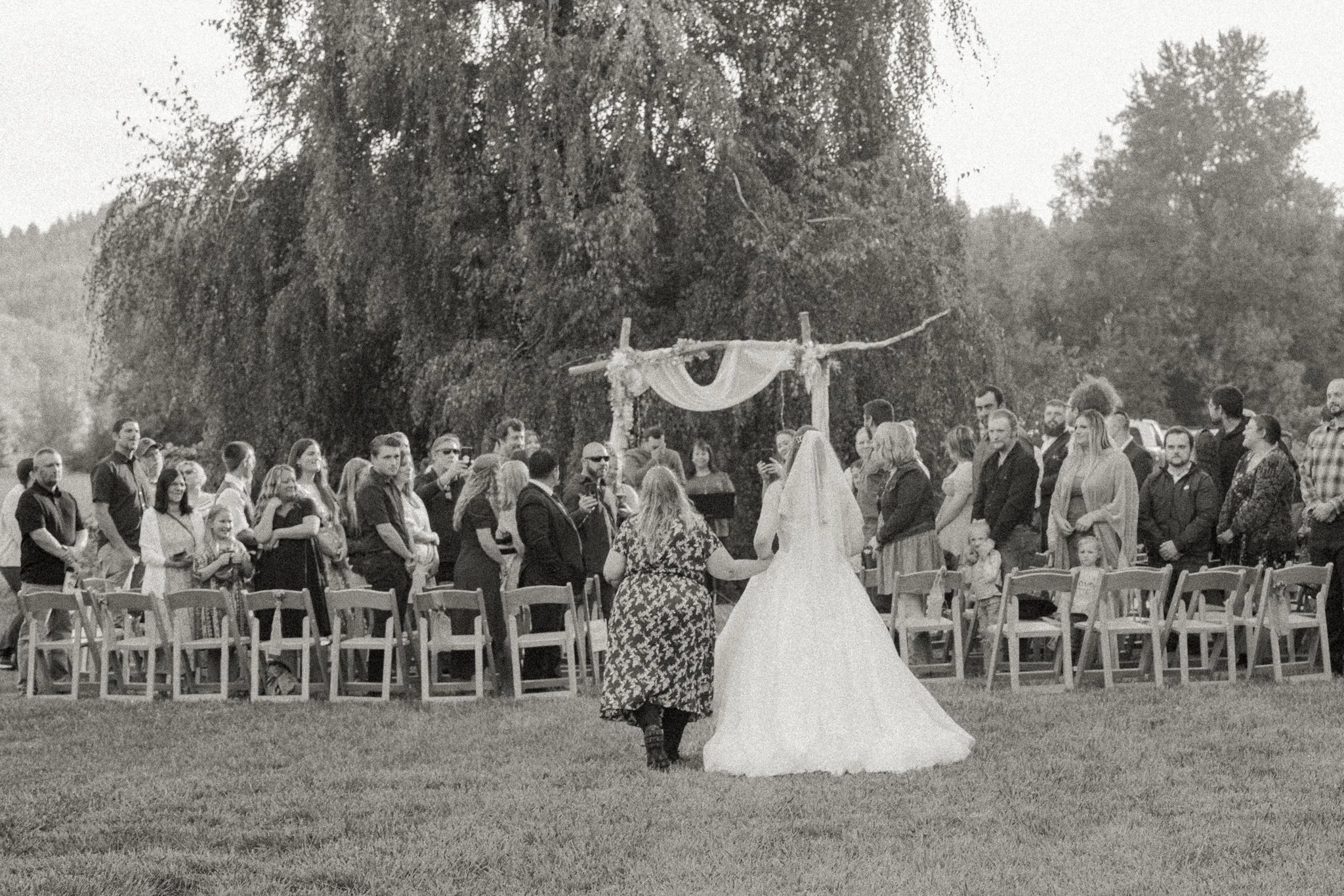 A black and white photo of a wedding ceremony outdoors with a gathering of guests around a bride and groom standing under an arch decorated with drapery and flowers, on a grassy area with trees in the background.