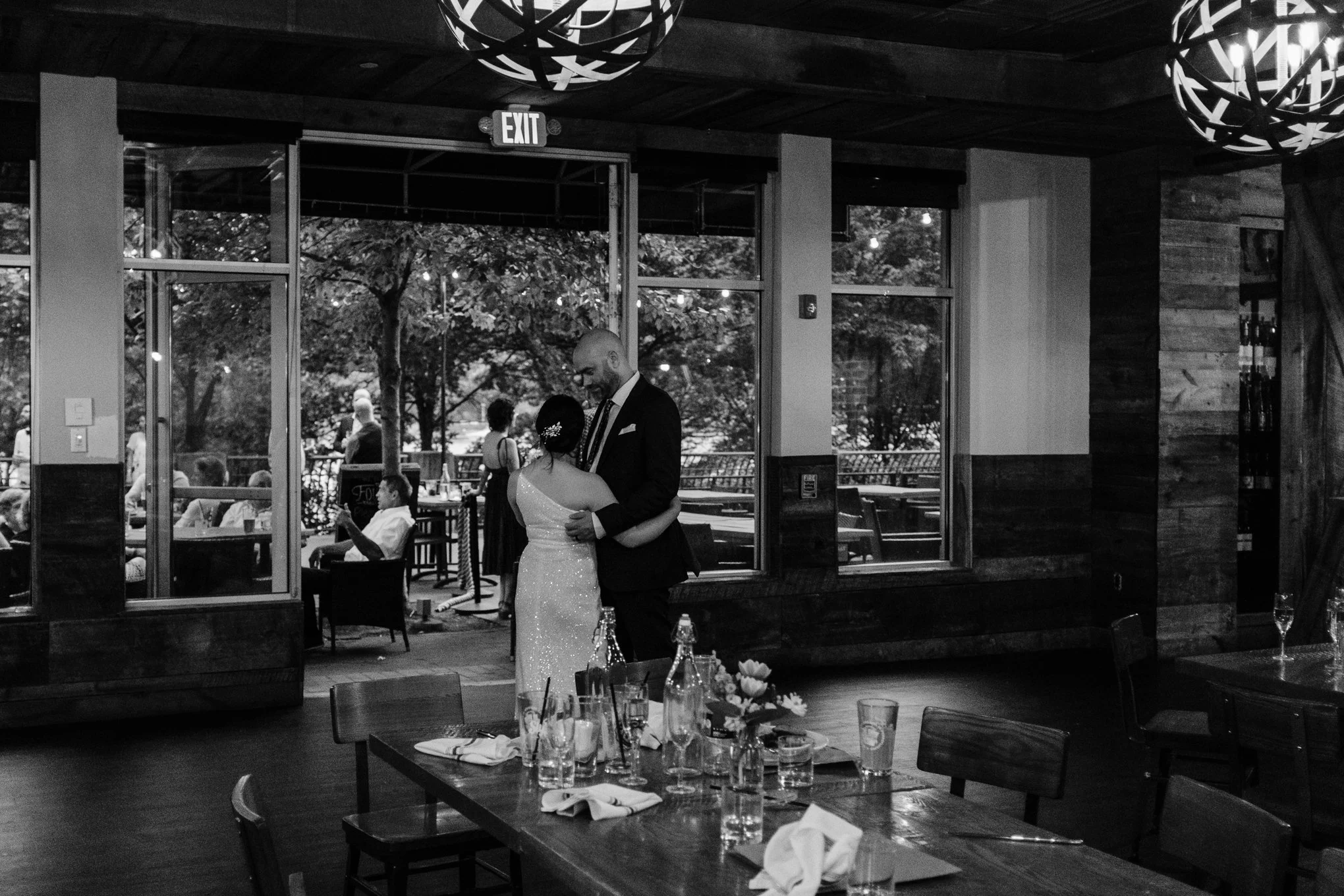 A black and white photo of a bride and groom dancing inside a restaurant or event space, with tables and chairs in the foreground and people sitting and standing outside visible through a large open doorway.