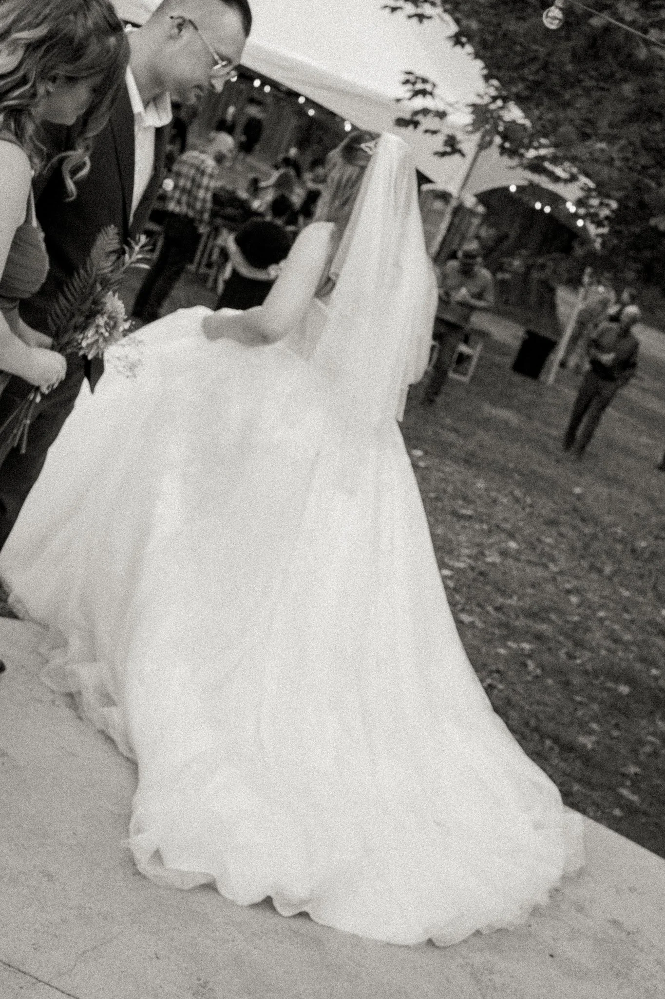 A bride in a white wedding gown and veil standing with groom in a suit during outdoor wedding ceremony, with guests and decorations visible in the background.