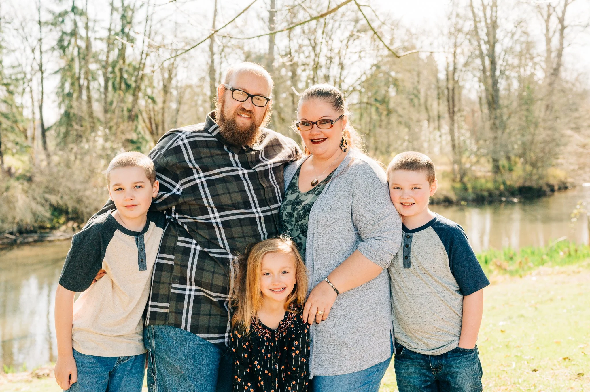A smiling family of six standing outdoors near a river, with trees in the background, on a sunny day.