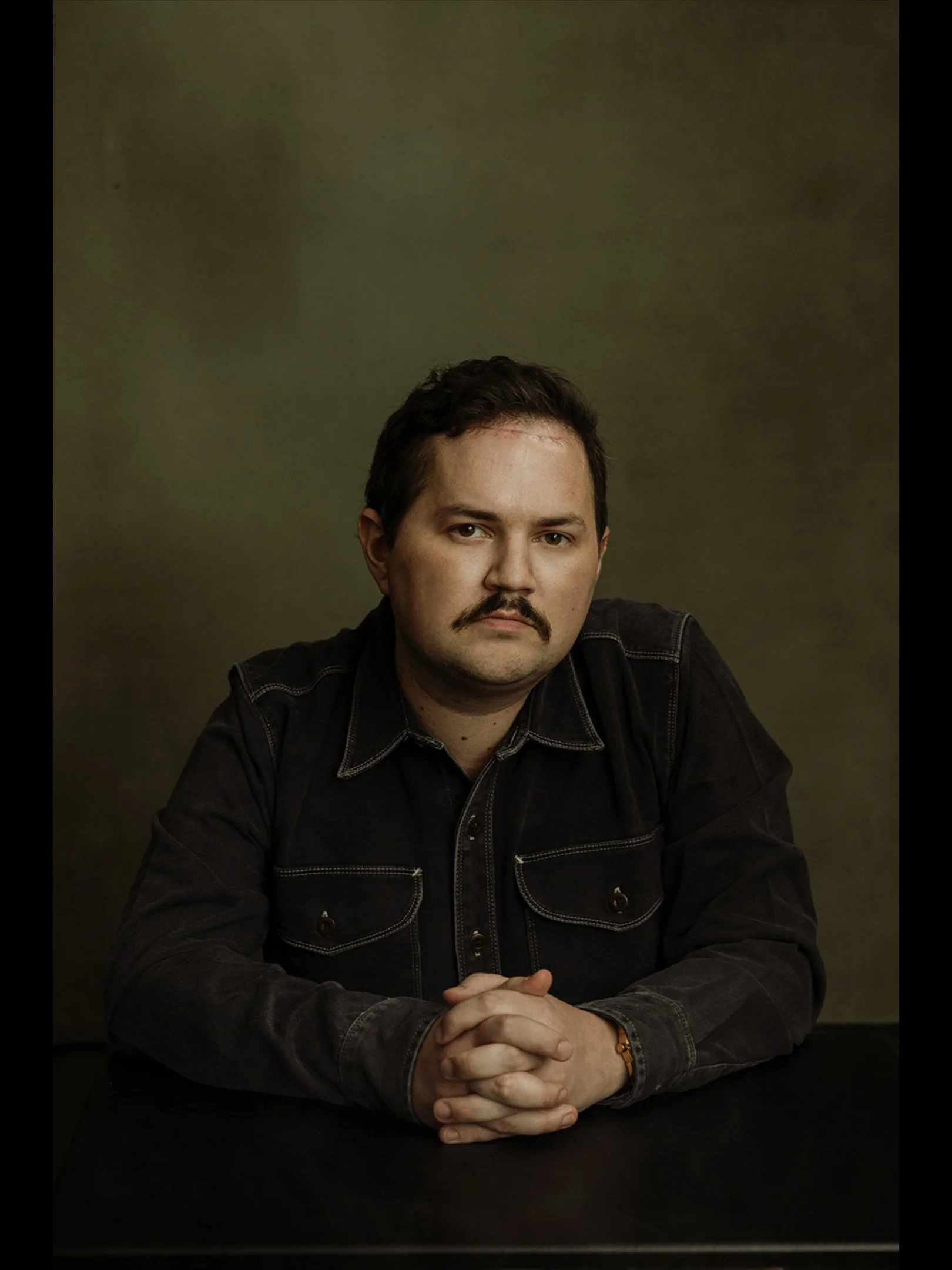 A man with dark hair and a mustache sitting at a table with hands clasped, wearing a black denim jacket, against a dark, textured green background.
