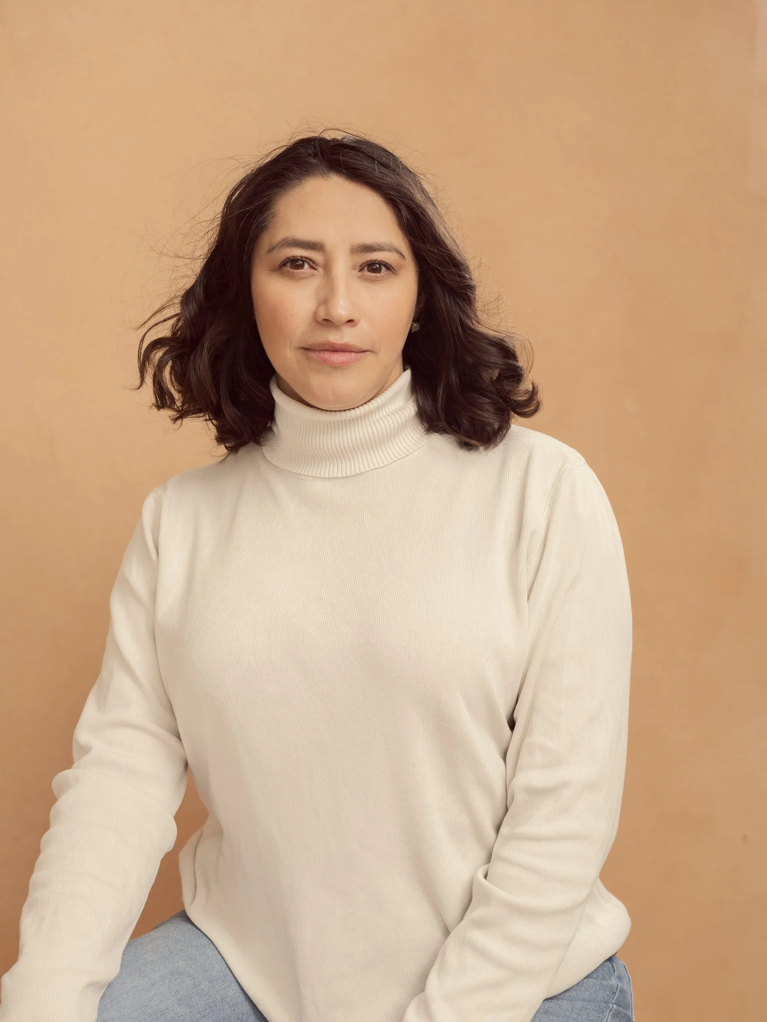 A woman with shoulder-length dark hair, wearing a cream turtleneck sweater, sitting against a light beige background.