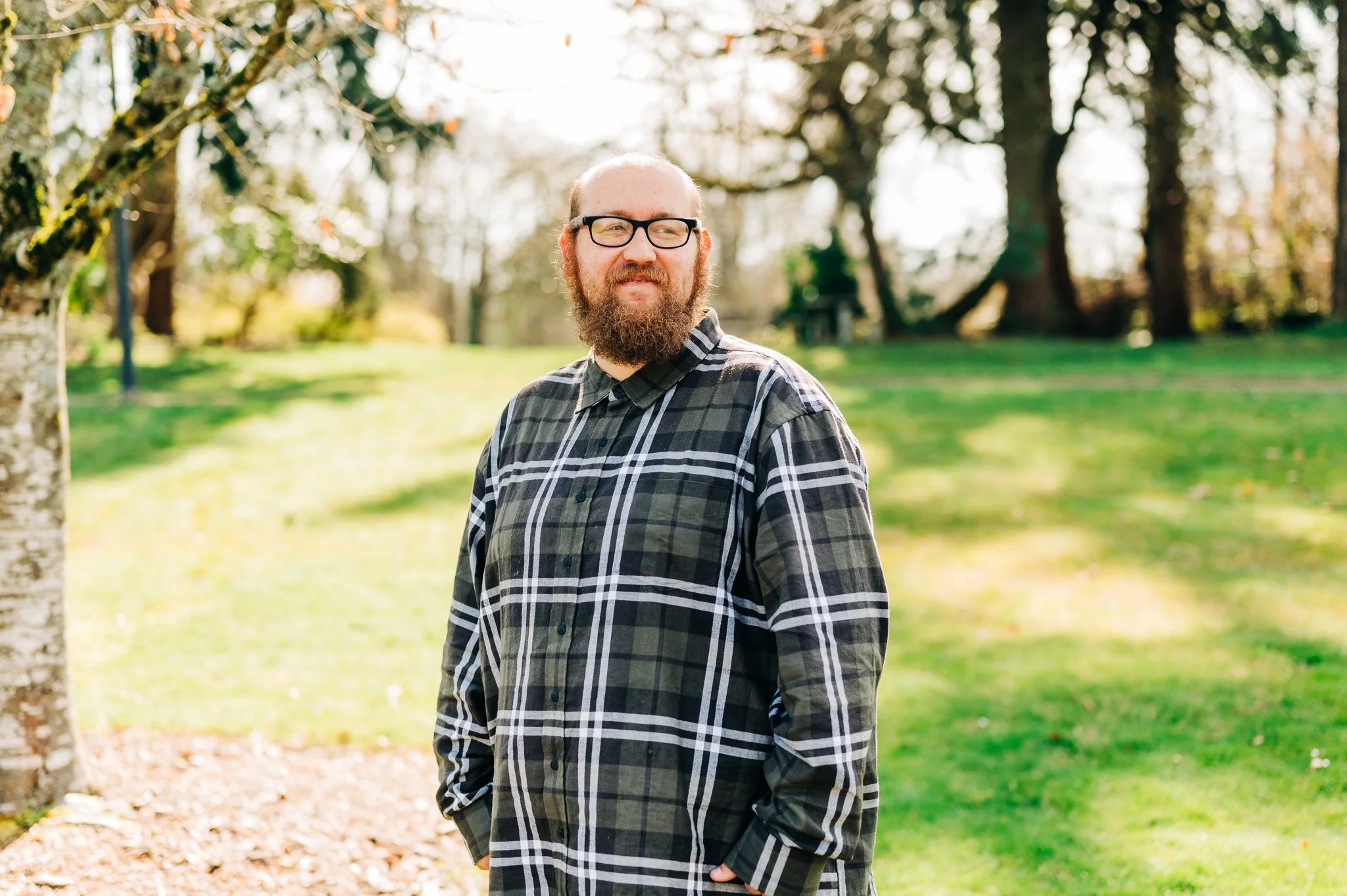A man with glasses and a beard standing outdoors on a sunny day, wearing a plaid shirt in a park with green grass and trees.