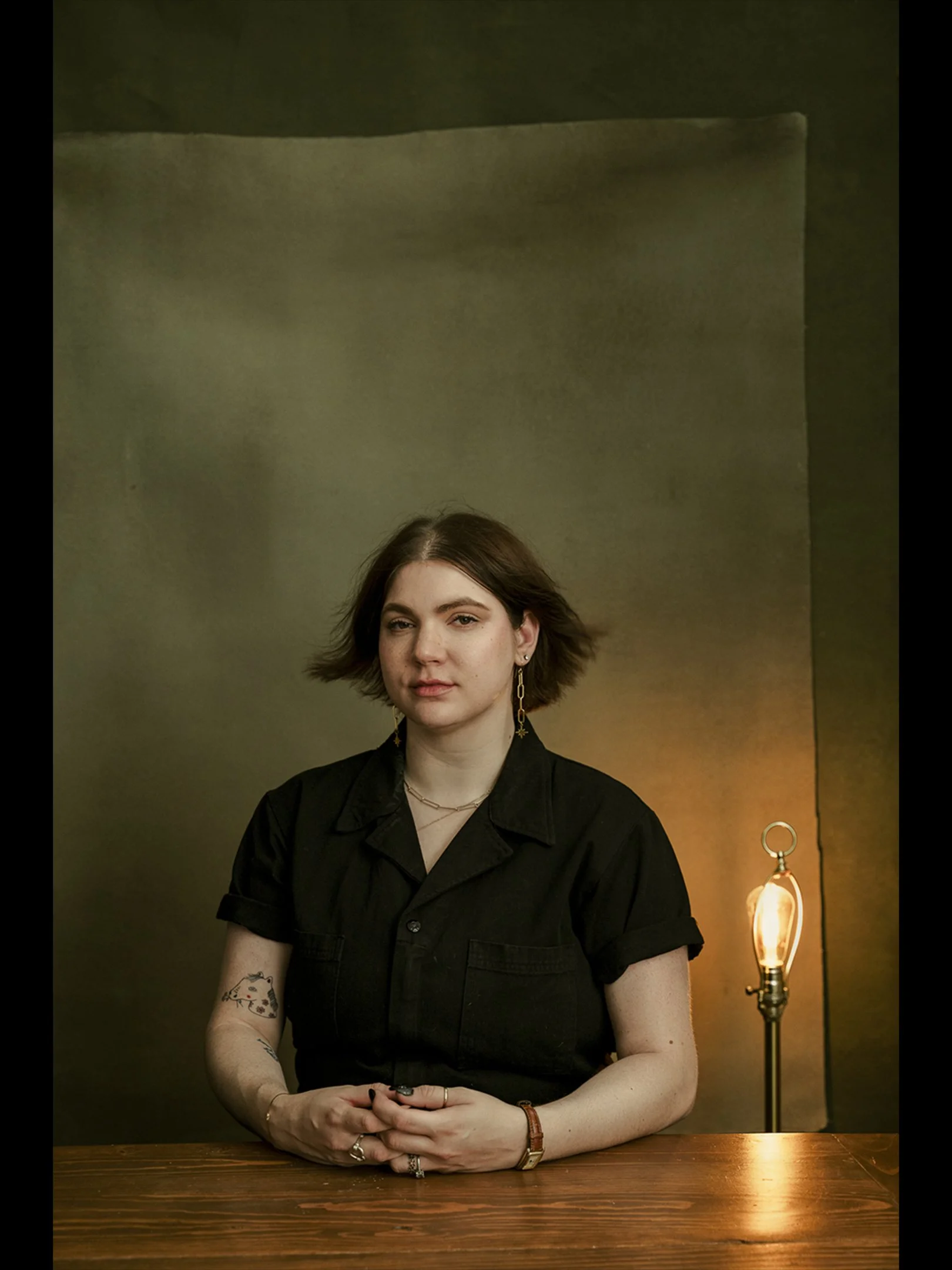 A woman with short brown hair wearing a black shirt and jewelry, sitting at a wooden table with a lamp in the background.