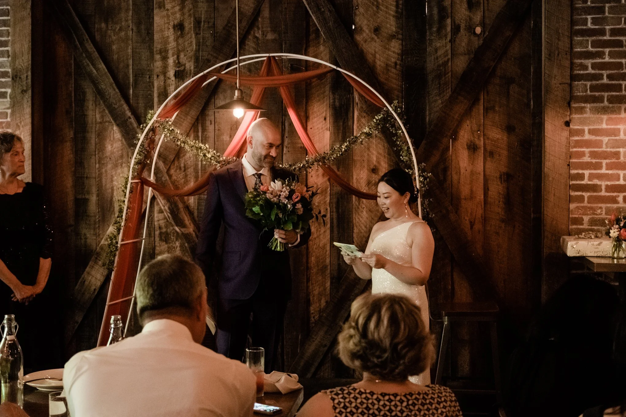 A wedding ceremony with a bride and groom exchanging vows under a decorative arch with draped fabric and greenery, inside a rustic venue with wooden walls and brick accents, with guests seated and watching.