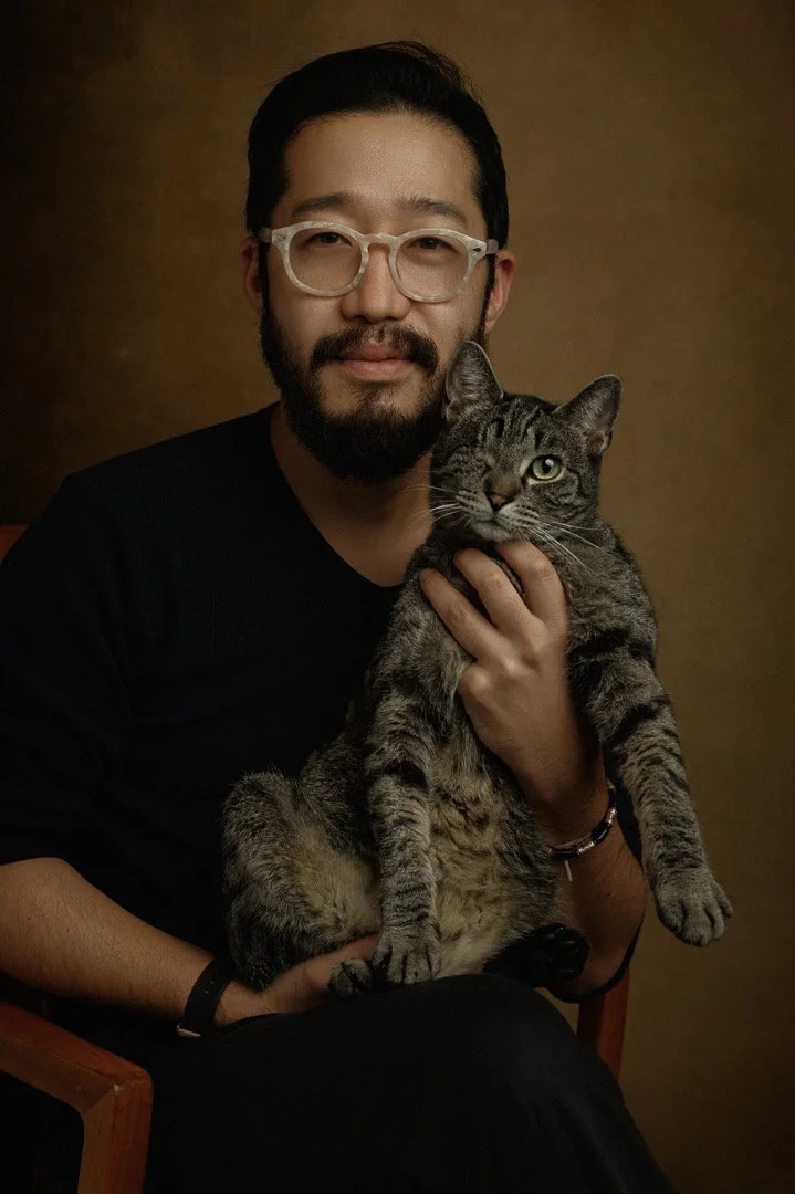 Portrait of a man with glasses holding a tabby cat, both looking at the camera against a brown background.