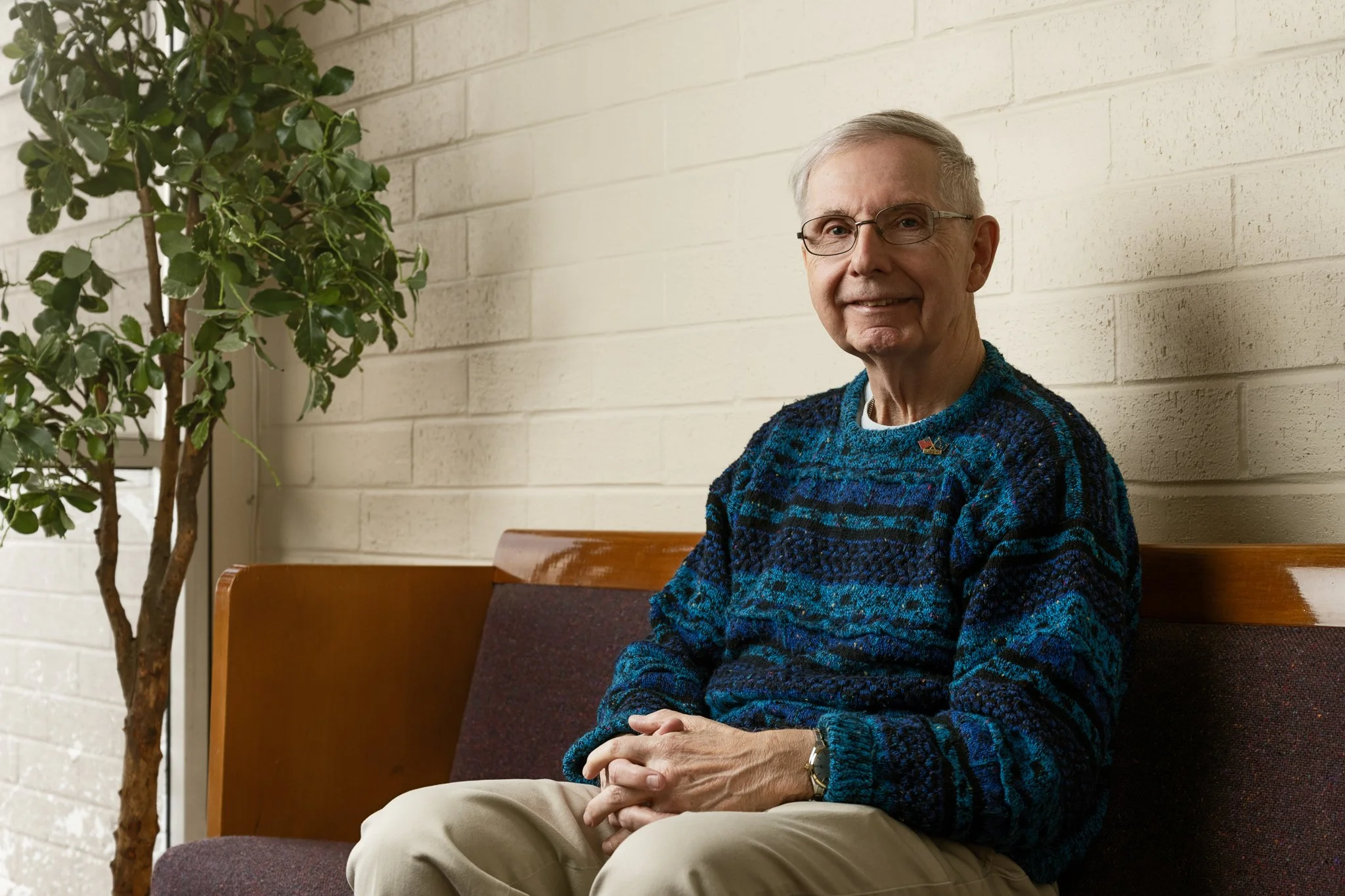 An elderly man with gray hair, glasses, and a friendly smile sitting on a wooden bench, wearing a blue patterned sweater and beige pants, beside a potted plant against a white brick wall.
