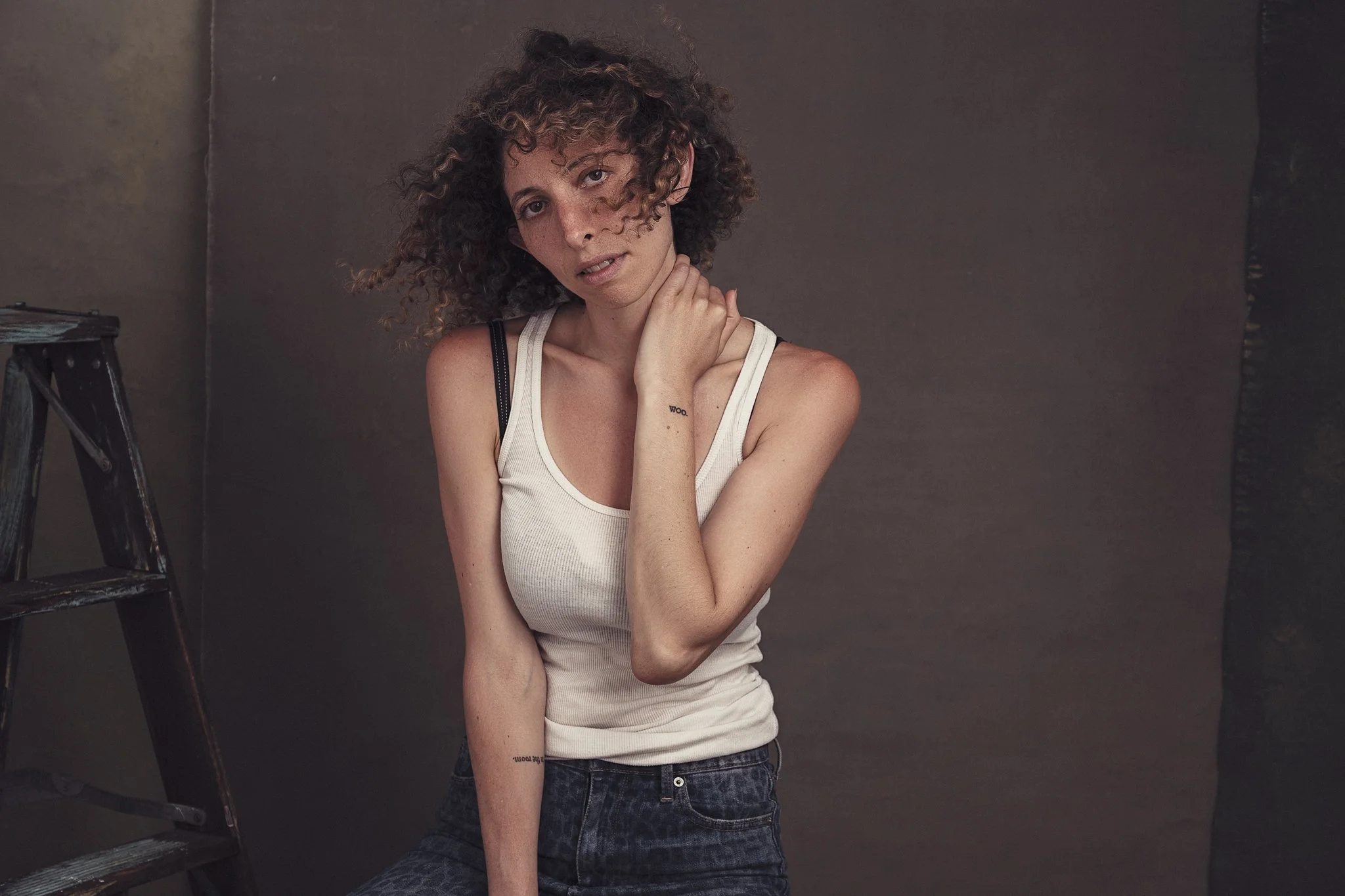 A woman with curly hair and freckles, wearing a white tank top and dark jeans, standing against a dark background next to a black stepladder, touching her neck and shoulder with a contemplative expression.