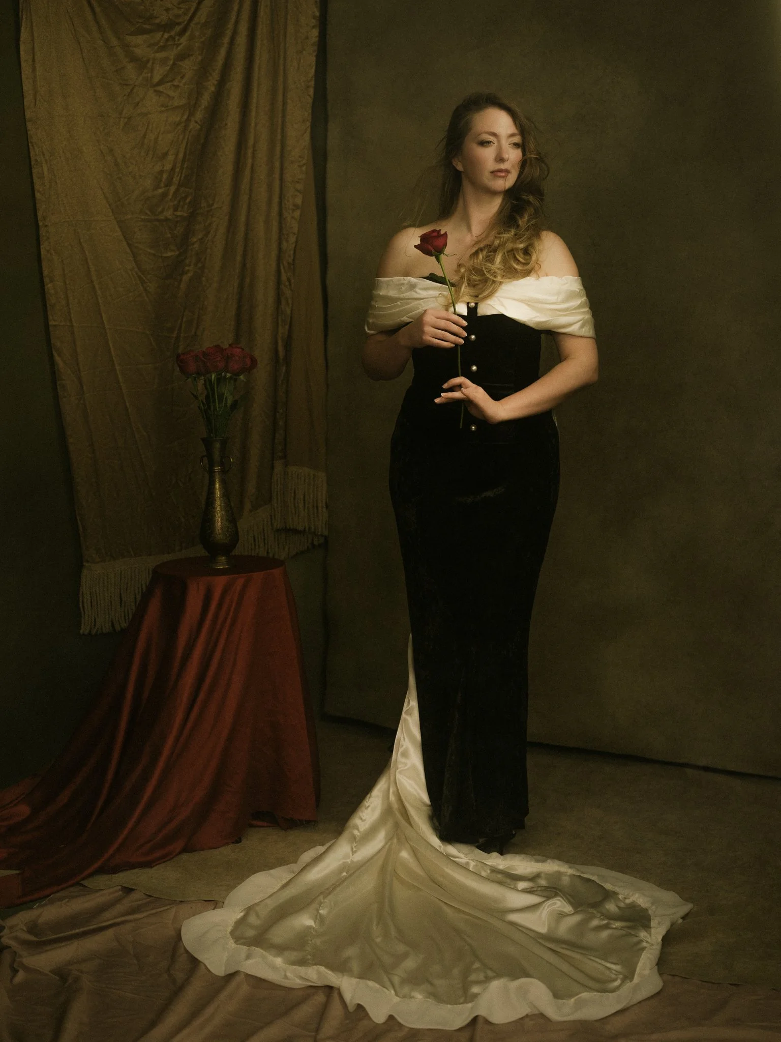 A woman standing, holding a single red rose, wearing a black and white gown with a train, on a neutral background with a tall vase of red roses on a table nearby.